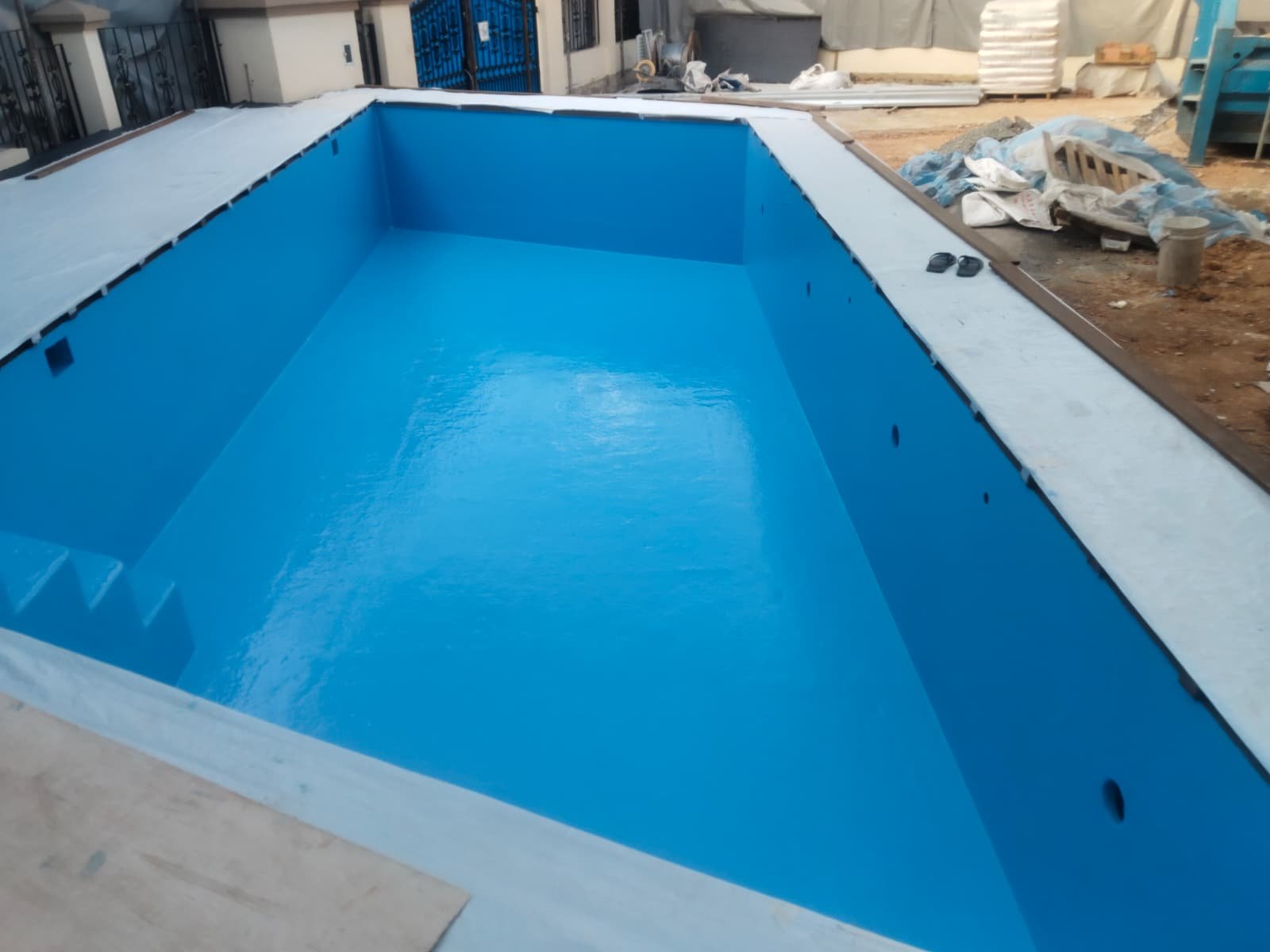 Empty swimming pool with a blue interior under construction, surrounded by construction materials and tools.