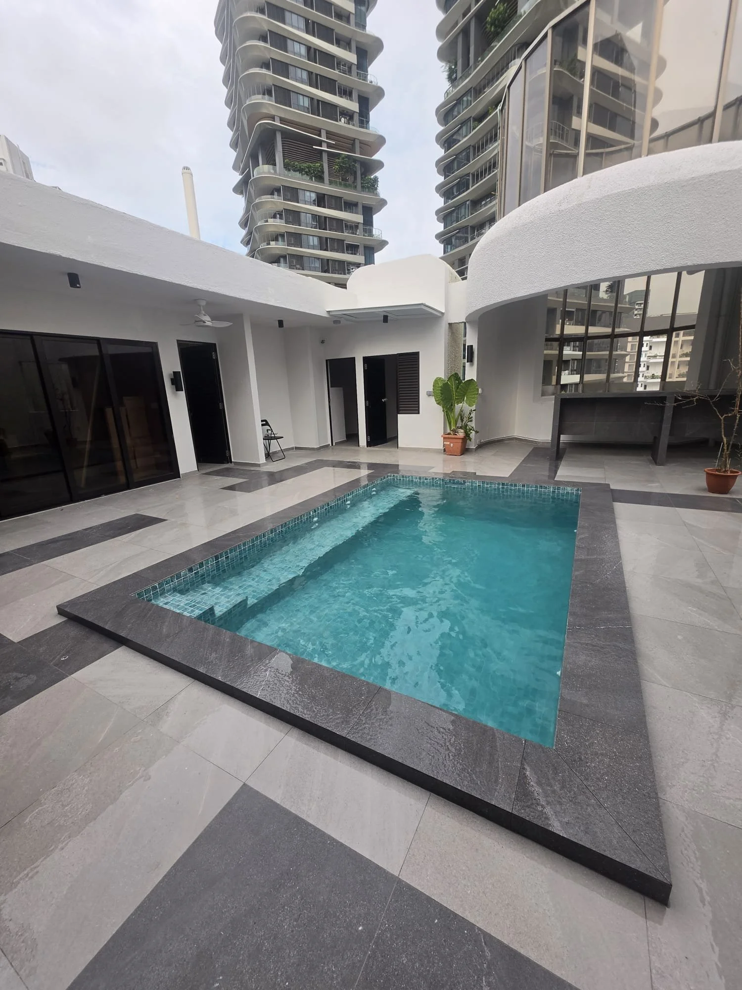 Outdoor swimming pool area in a modern apartment complex with high-rise buildings in the background, potted plants, glass doors, black chairs, and a canopy.