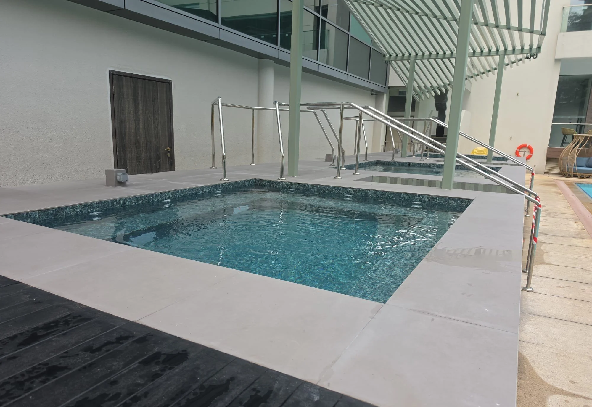 Modern apartment building outdoor pool area with small rectangular pool, metal railings, a covered patio with slatted roof, glass balconies, and a wooden fence with seating nearby.