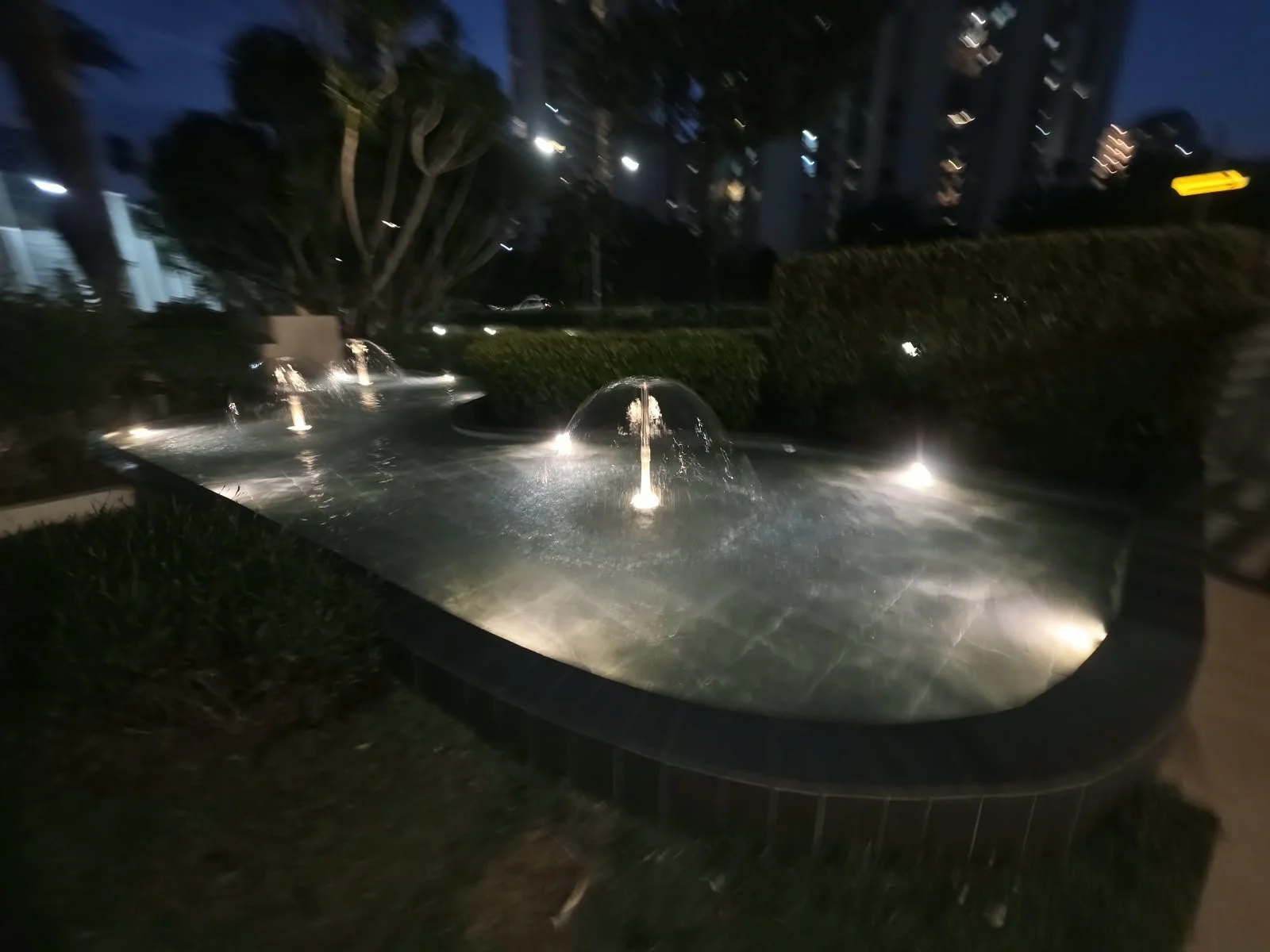 Nighttime view of a decorative water fountain with illuminated water jets in an urban park, surrounded by bushes and trees.
