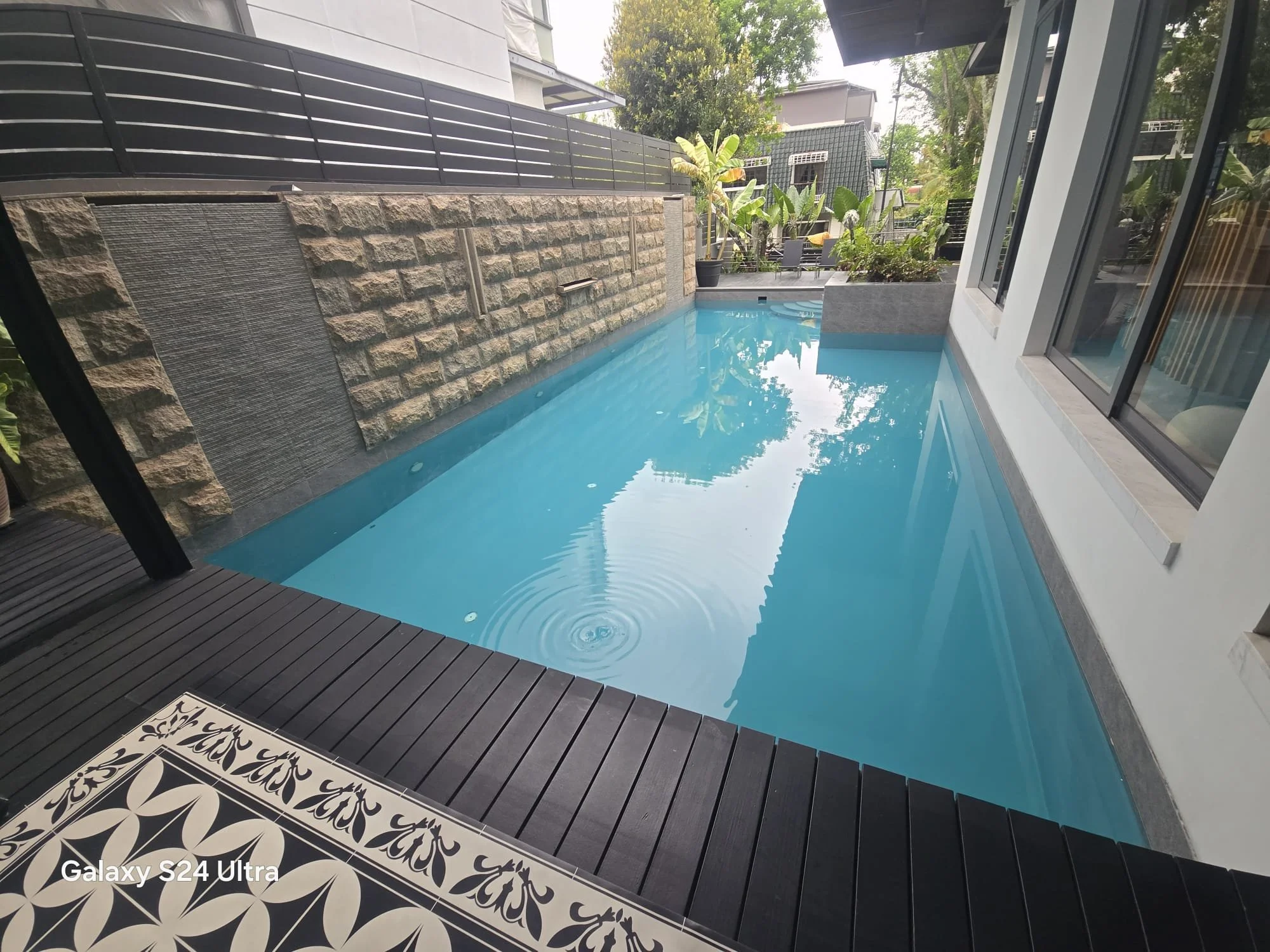 A backyard with a small pool, surrounded by decorative tiles and black wood decking. In the background, there are lush green plants and trees, a seating area with chairs, and modern black fencing, with neighboring buildings and trees visible beyond.