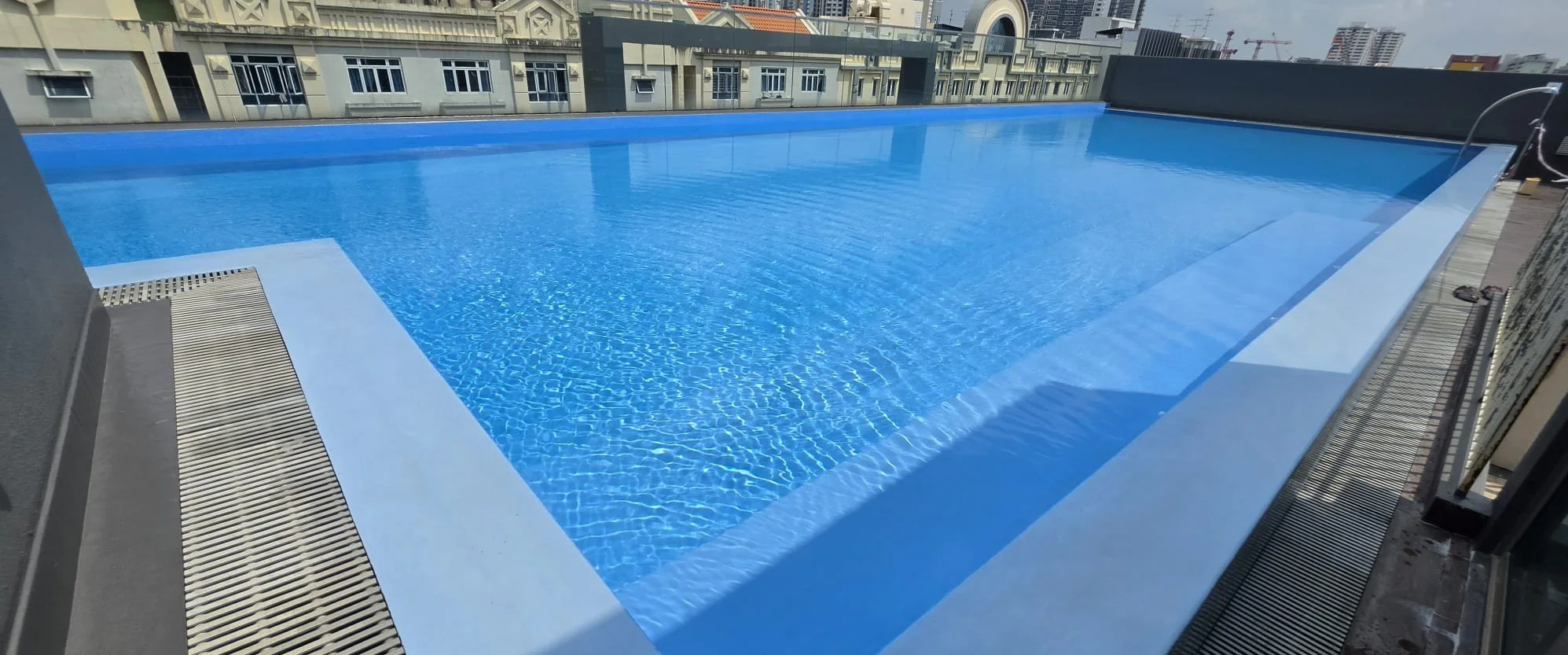 A rooftop swimming pool with clear blue water, surrounded by a gray deck and city buildings in the background.