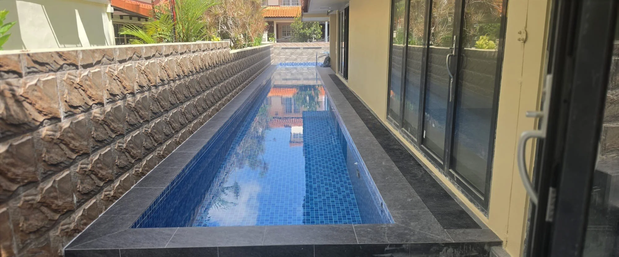 A narrow outdoor pool with blue mosaic tiles on the floor, bordered by black tiles, running alongside a building with large glass sliding doors. There's a stone wall with plants on the left side and a house with a red-tiled roof visible in the background.
