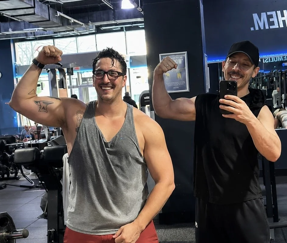 Two men posing in a gym, flexing their biceps and smiling at the camera. One is wearing a gray tank top and glasses, while the other is in a black sleeveless hoodie and a cap.