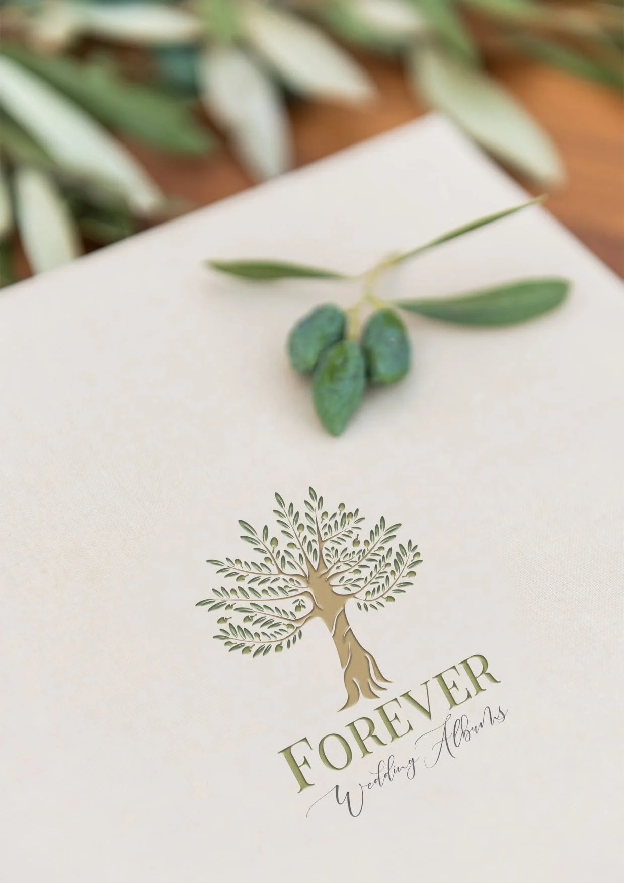 A wedding album with a white cover featuring a gold-embossed tree logo and the text "Forever" and "Wedding Albums." A small branch with green leaves rests on top of the album.