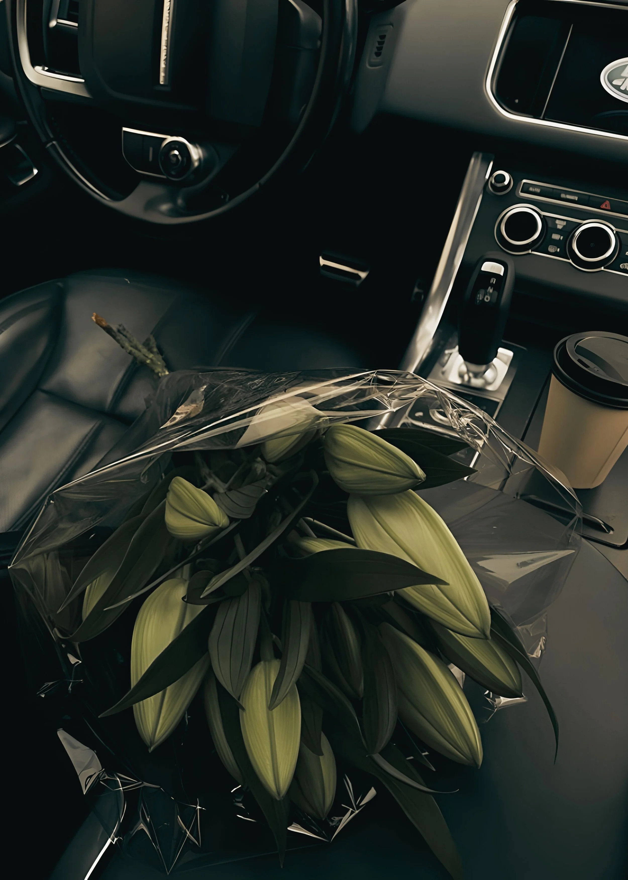 Inside of a car with the dashboard, steering wheel, and gear shift visible. There is a bouquet of greenish flowers wrapped in clear plastic on the passenger seat.