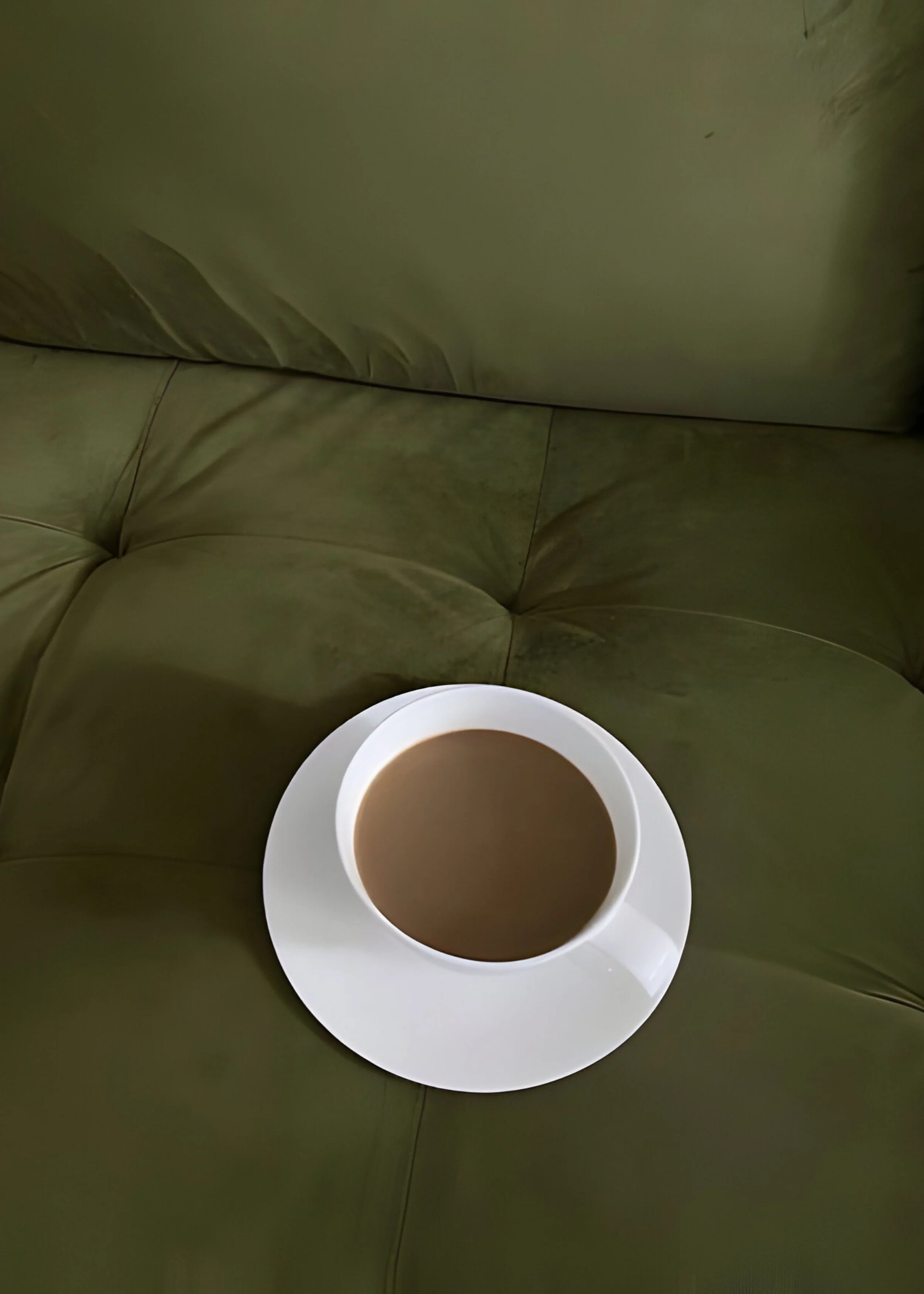 A cup of coffee on a white saucer placed on an olive green sofa.