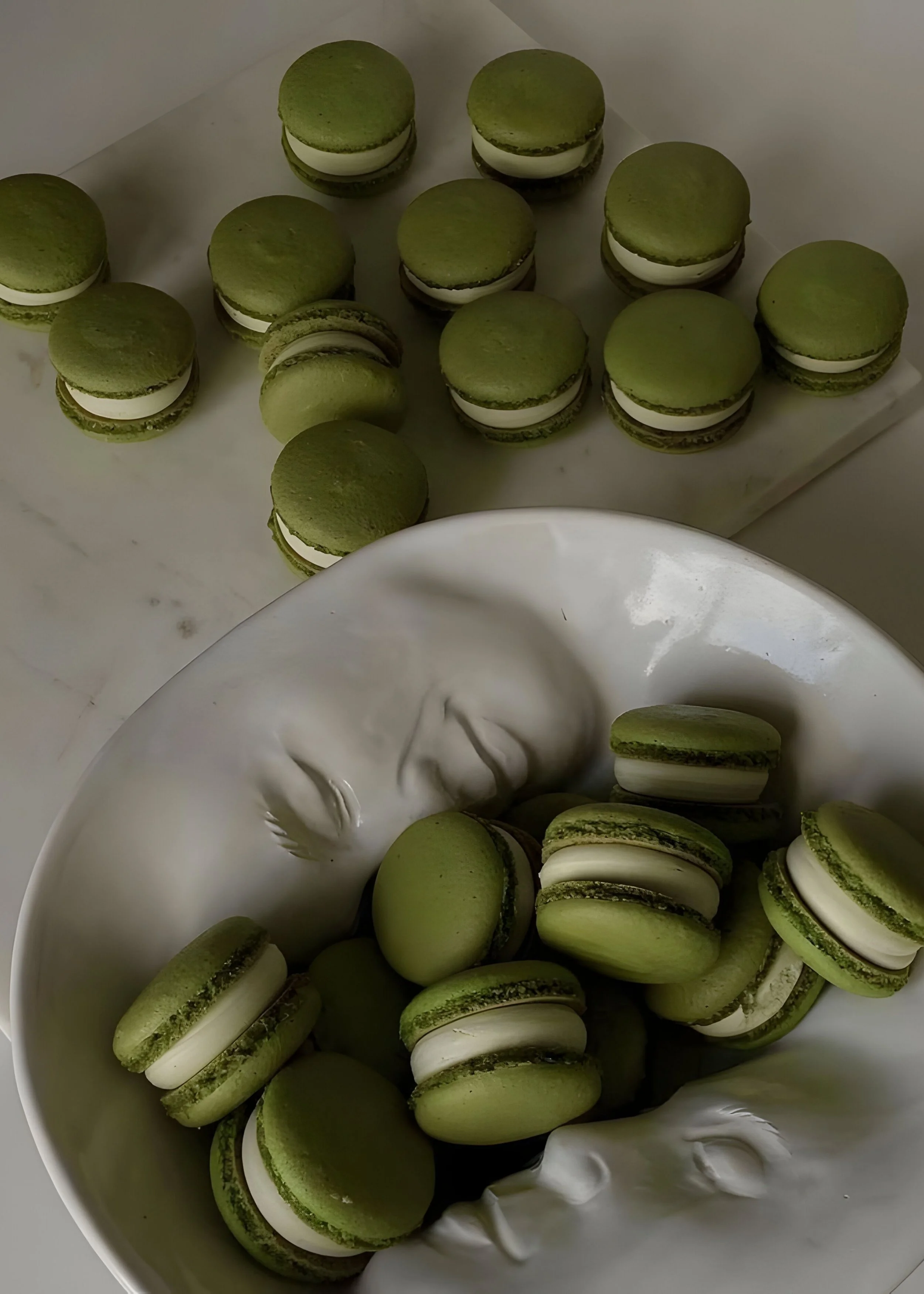 Green macarons with white filling arranged on a white porcelain dish and a marble surface.