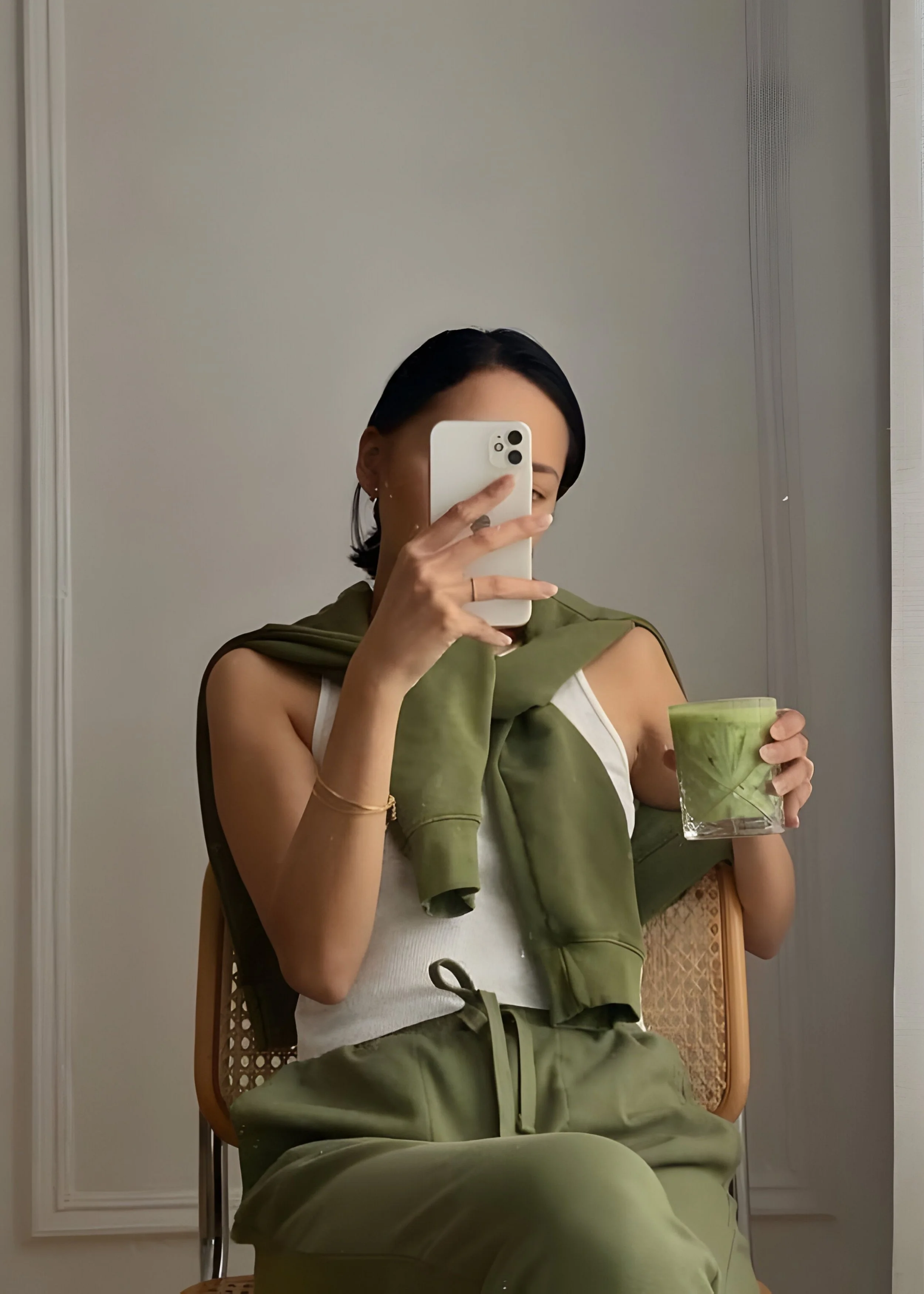 Woman taking a mirror selfie with a smartphone, sitting on a wooden chair, holding a green drink in a glass.
