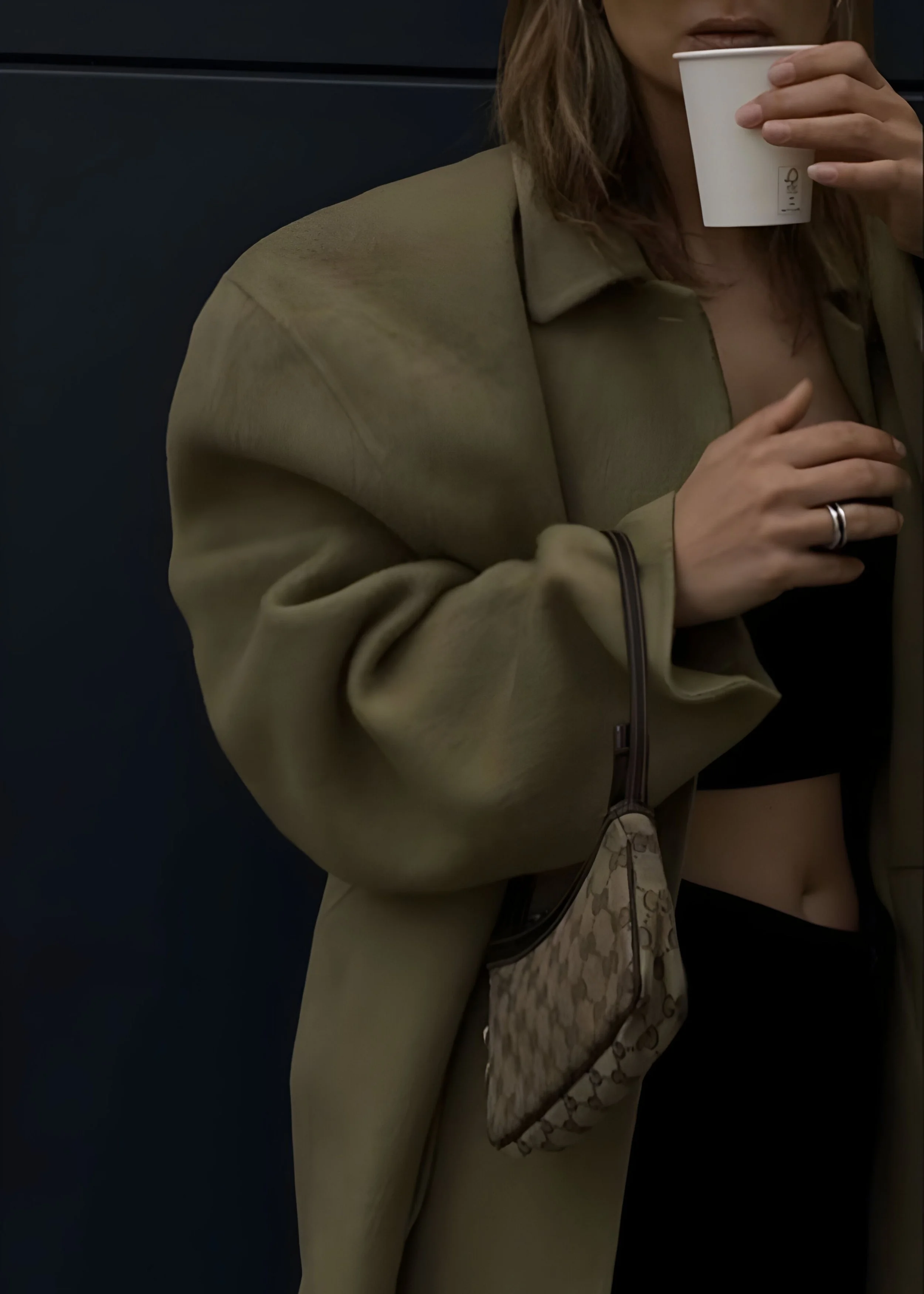 A woman with brown hair wearing a beige jacket, holding a white paper cup.