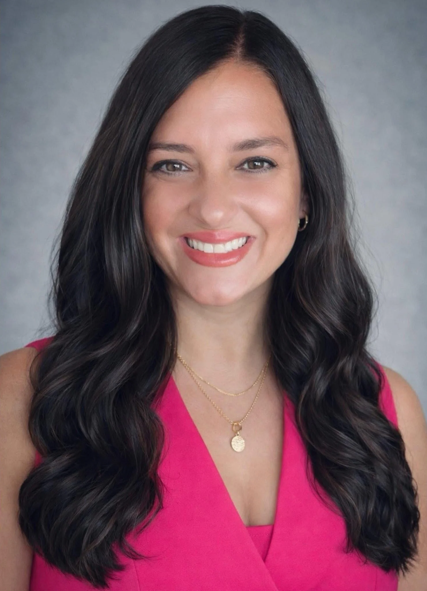 Close-up portrait of a woman with long dark wavy hair, wearing a pink sleeveless top, gold jewelry, smiling against a neutral background.