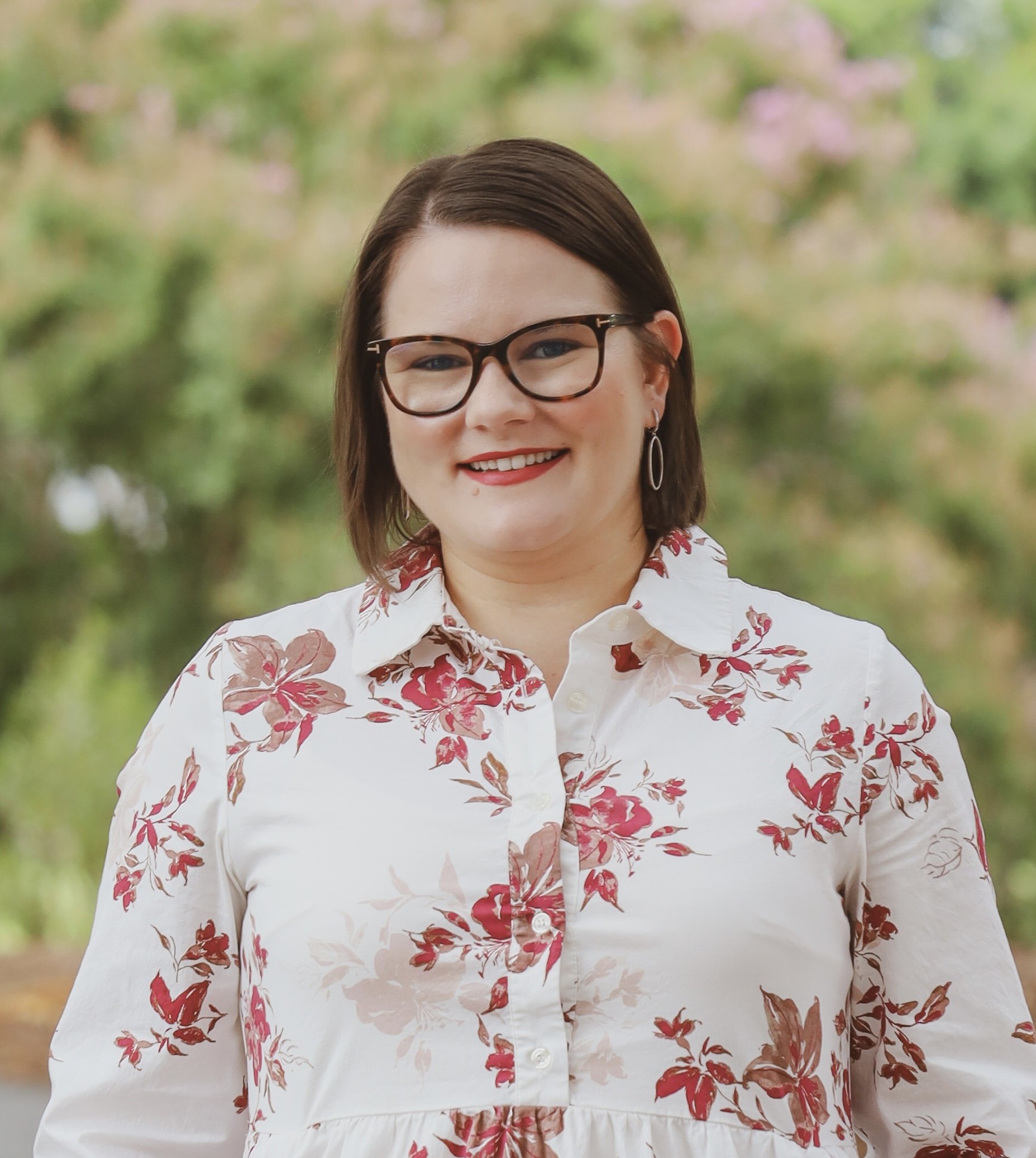 Smiling woman with dark brown hair, wearing glasses and earrings, in a floral white blouse, outdoors with green and pink blurred background.