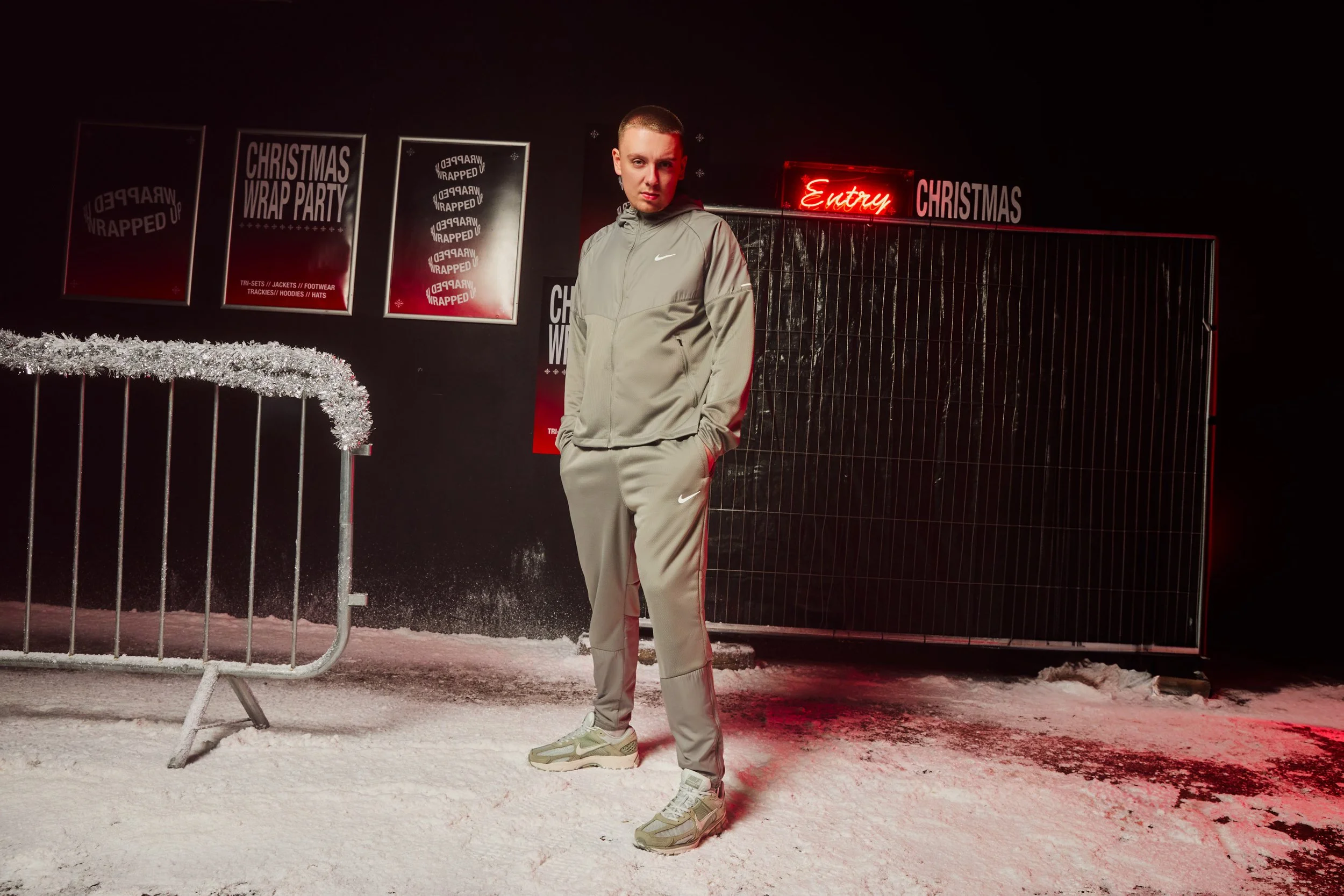 Aitch the music Artist standing in front of a black wall with Christmas decorations, signs reading 'Christmas Wrap Party' and 'Entry,' wearing a light gray Nike tracksuit and sneakers, on snowy ground at night. For Nick Gee Productions