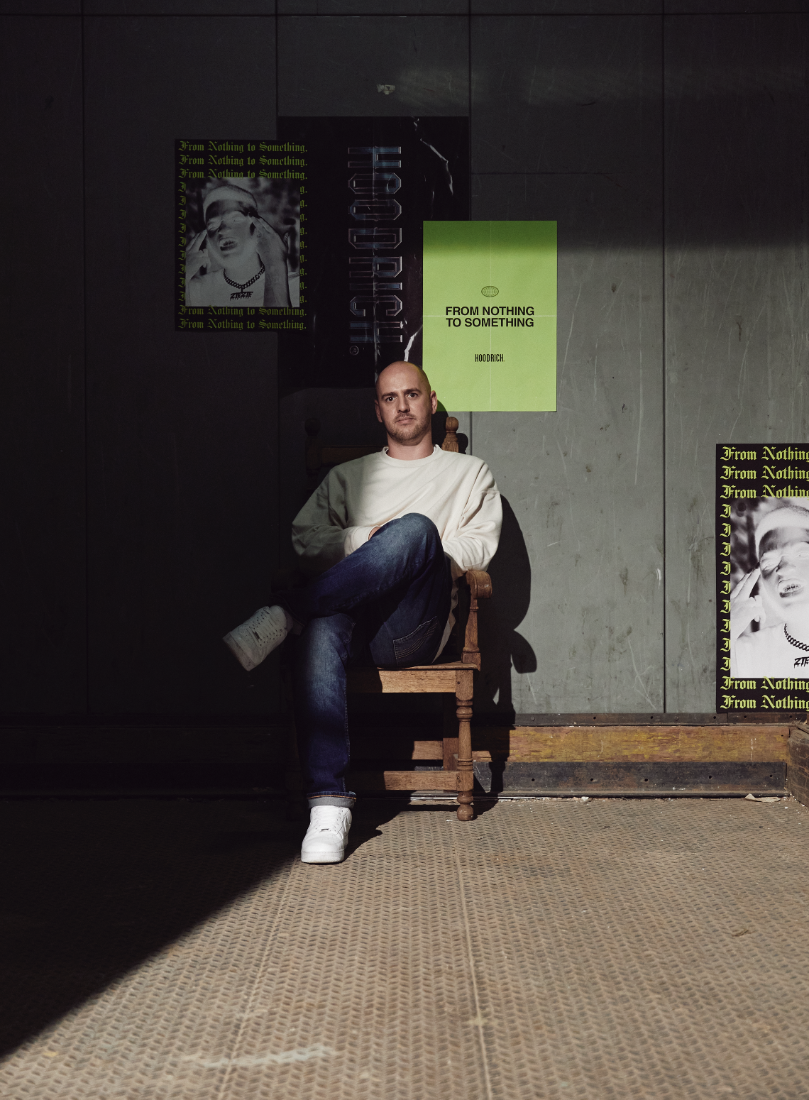 A man sitting on a wooden chair against a gray wall with posters reads 'From Nothing to Something' and 'Hoodrich.' He wears a white sweatshirt, blue jeans, and white sneakers, with one leg crossed over the other. Shadows cast on the wall emphasize the scene's lighting.