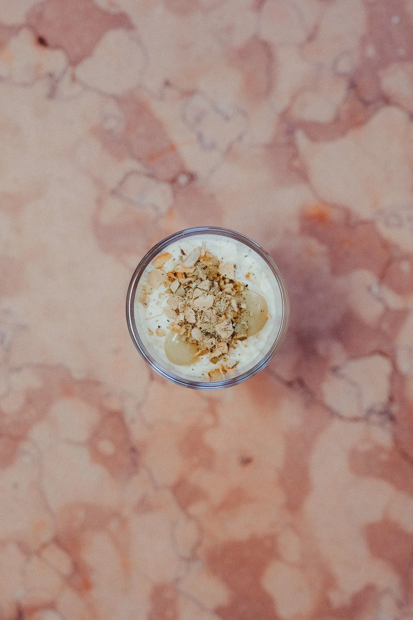 Top-down view of a cup of yogurt topped with chopped nuts, placed on a pink marble surface.