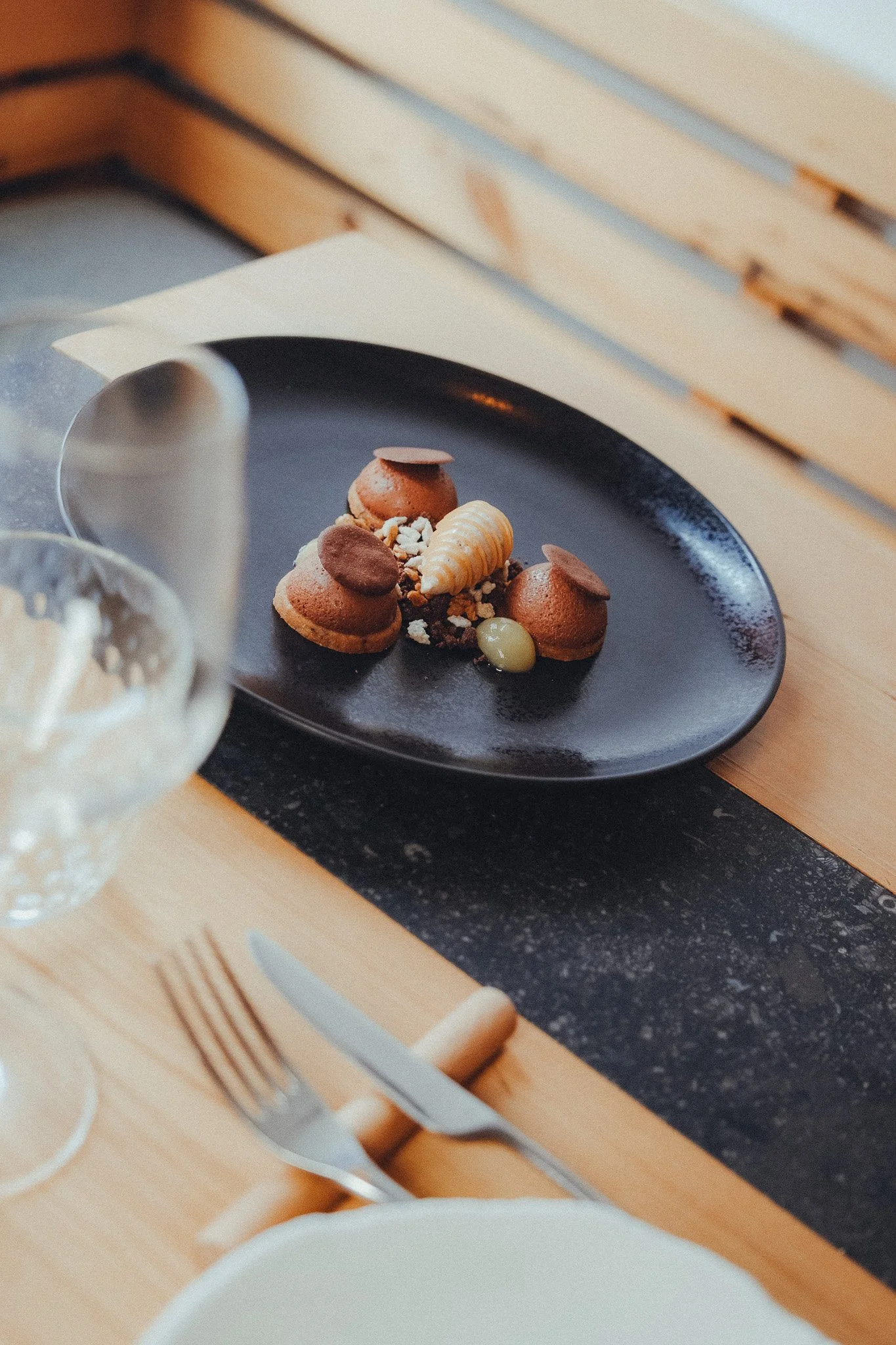 A black plate with assorted desserts including a seashell-shaped pastry, small round pastries with chocolate toppings, and a yellow-green bean-shaped candy, set on a wooden table near a glass of white wine.