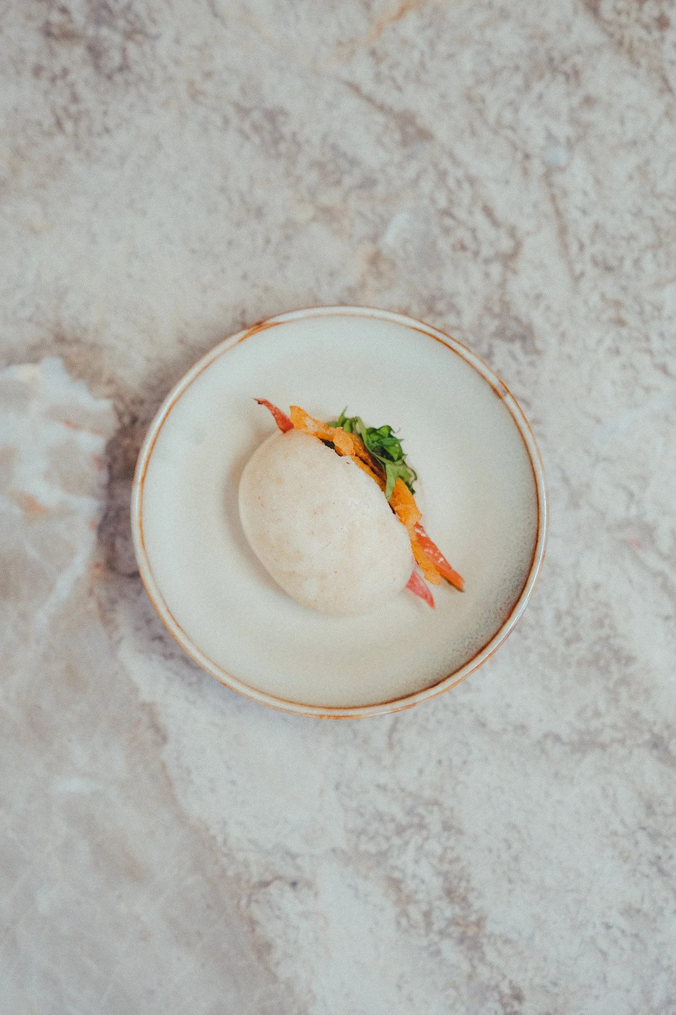 A white bao bun sandwich filled with crispy bacon, shredded carrots, and fresh greens on a small white plate.