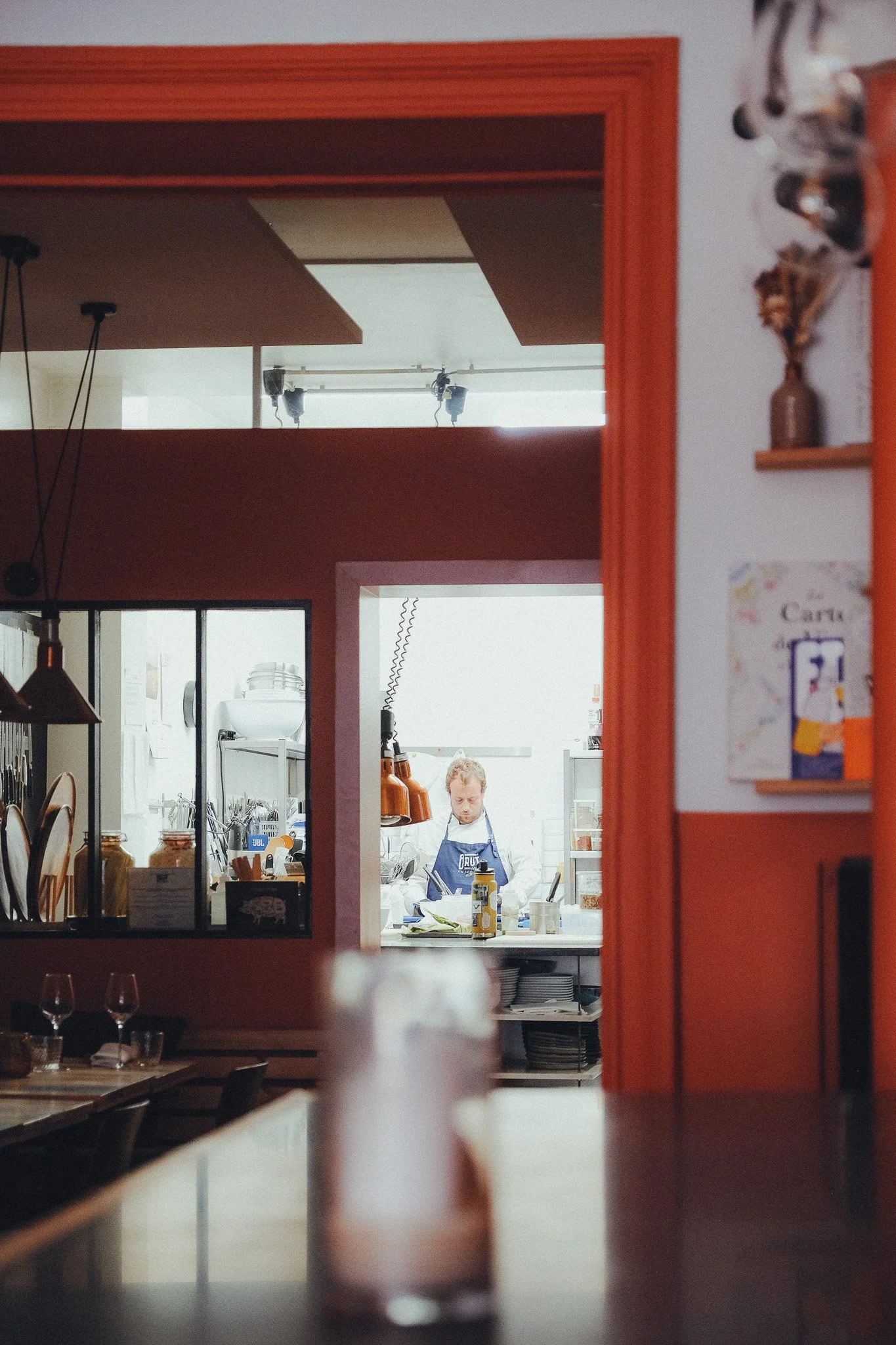 A person working in a bright kitchen seen through a window from a dining area with tables and wine glasses.