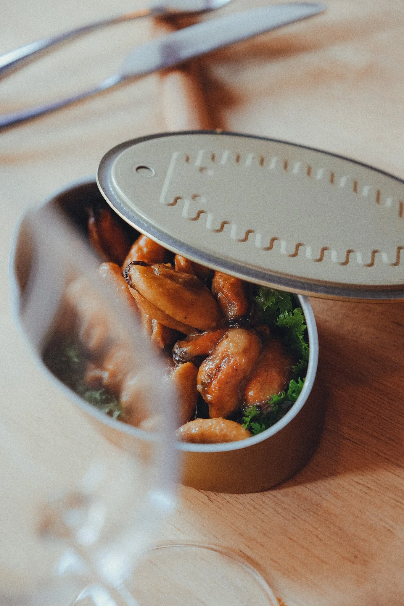 Close-up of a tin container with cooked chicken pieces garnished with fresh cilantro, partially covered by a metal lid, on a wooden surface. In the background, there is a fork and knife.