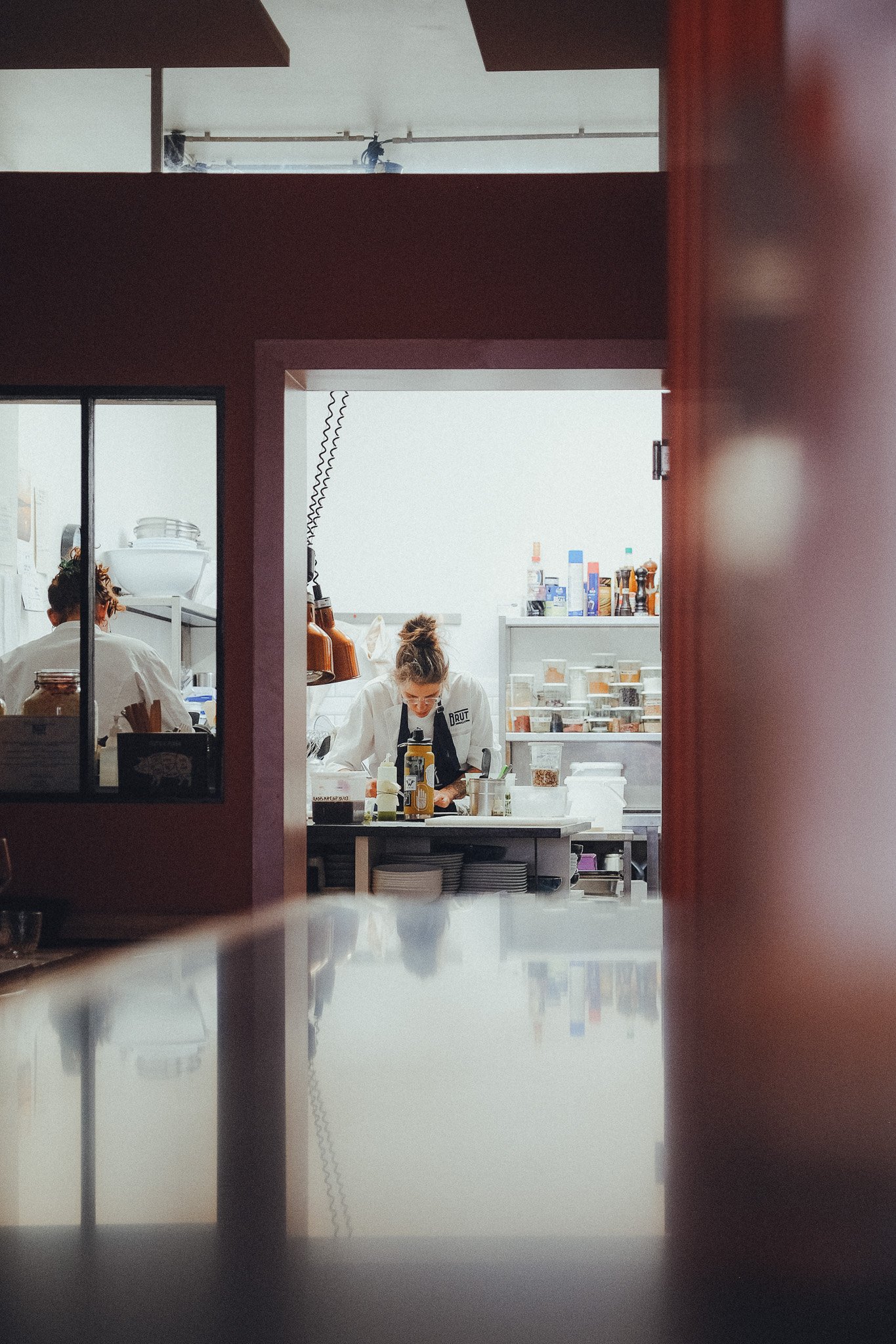 View of a busy kitchen seen through a window or opening, with a person working at a counter, shelves with various bottles and containers, and kitchen equipment.