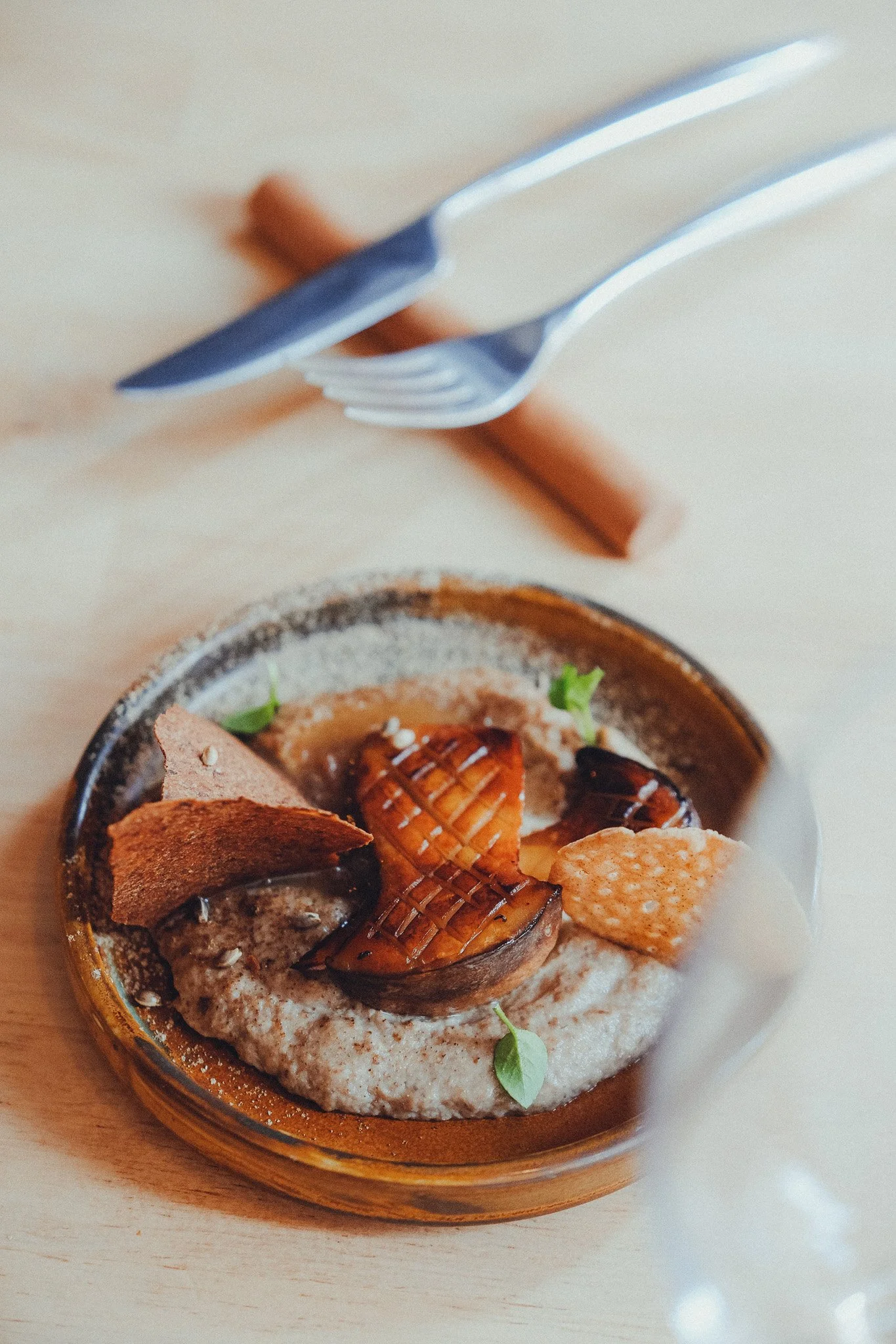 Close-up of a bowl of hummus topped with roasted vegetables, herbs, and crispy chips on a wooden surface.