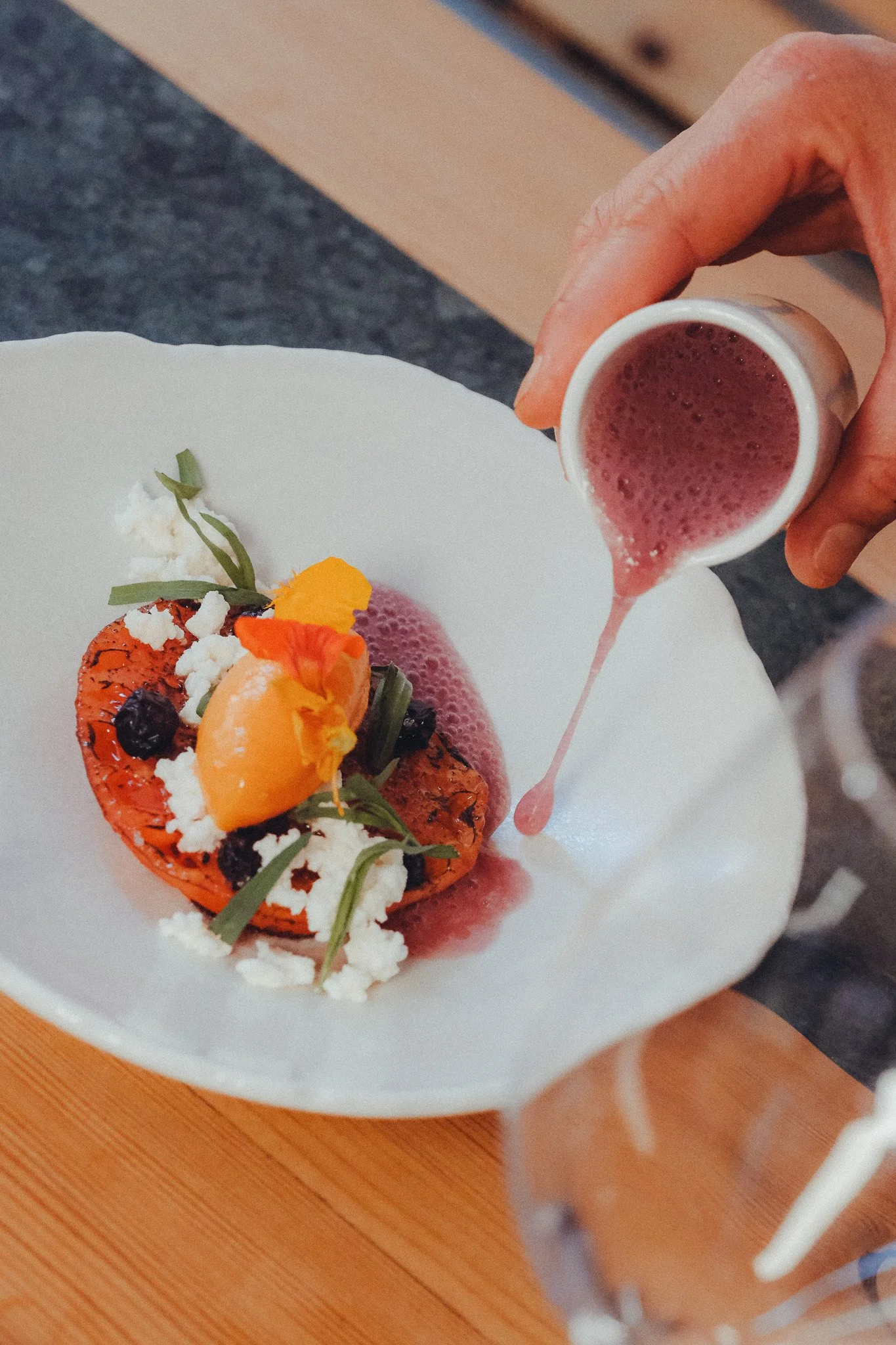 A dessert topped with whipped cream, fresh berries, and edible flowers on a white plate, with pink syrup being poured over it from a small pitcher.