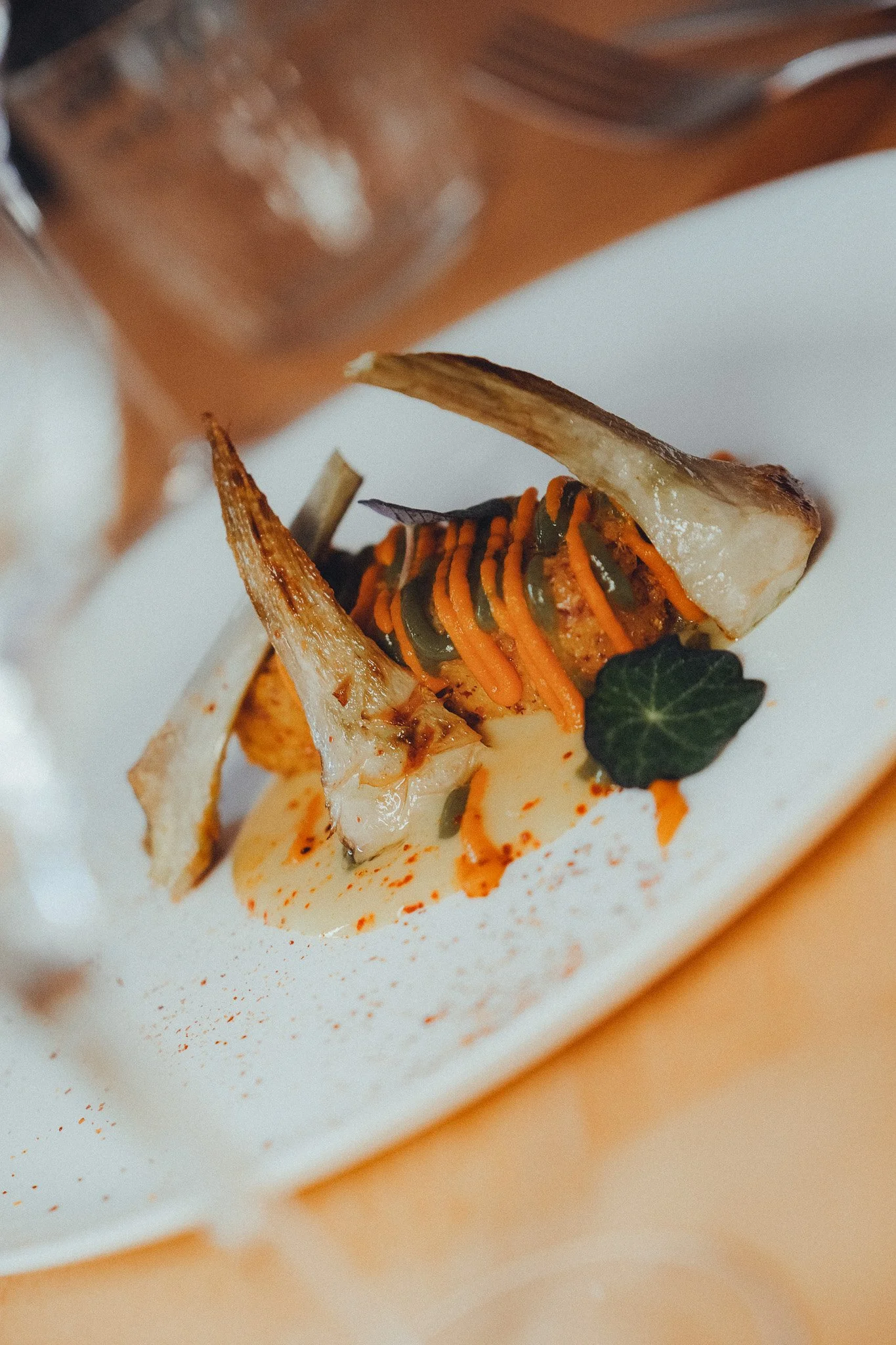 Close-up of a plated fish fillet garnished with shredded carrots, green herbs, and a drizzle of sauce on a white plate with a rim of red spice, on a wooden table.