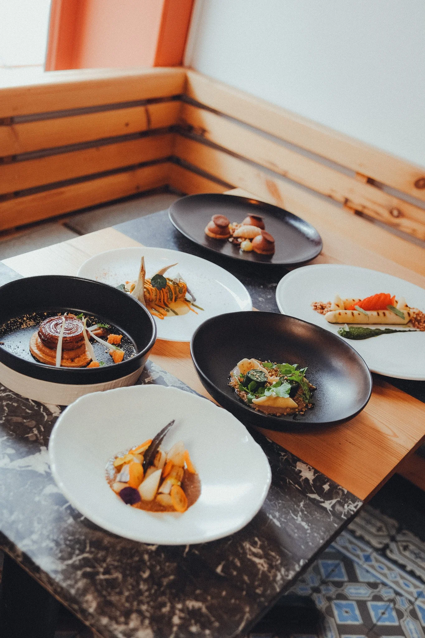 Assorted gourmet dishes on black, white, and ceramic plates on a marble table, with a wooden bench in the background.