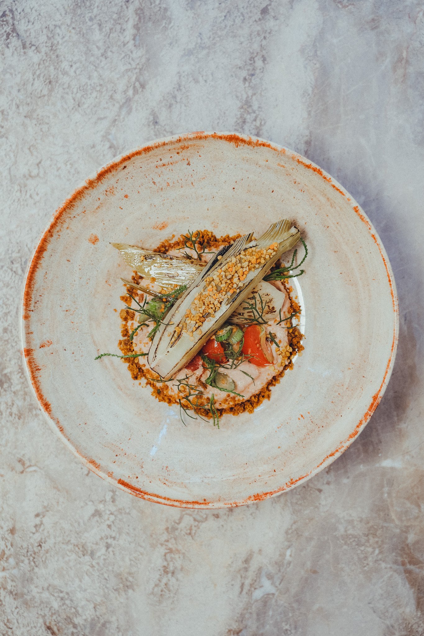 A white ceramic plate with an orange rim containing a fine, grainy mixture, garnished with roasted vegetables, herbs, and crunchy toppings.
