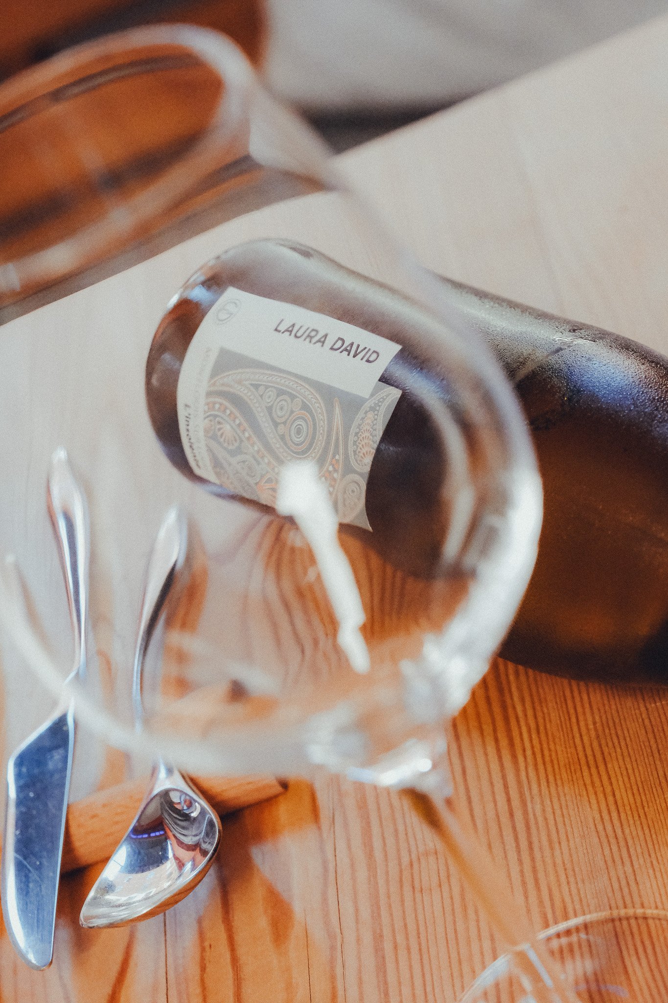 A clear glass mug with a white label reading 'Laura David' and an intricate design behind the text, sitting on a wooden table with a teaspoon and a metal spoon beside it. A dark bottle is also visible in the background.