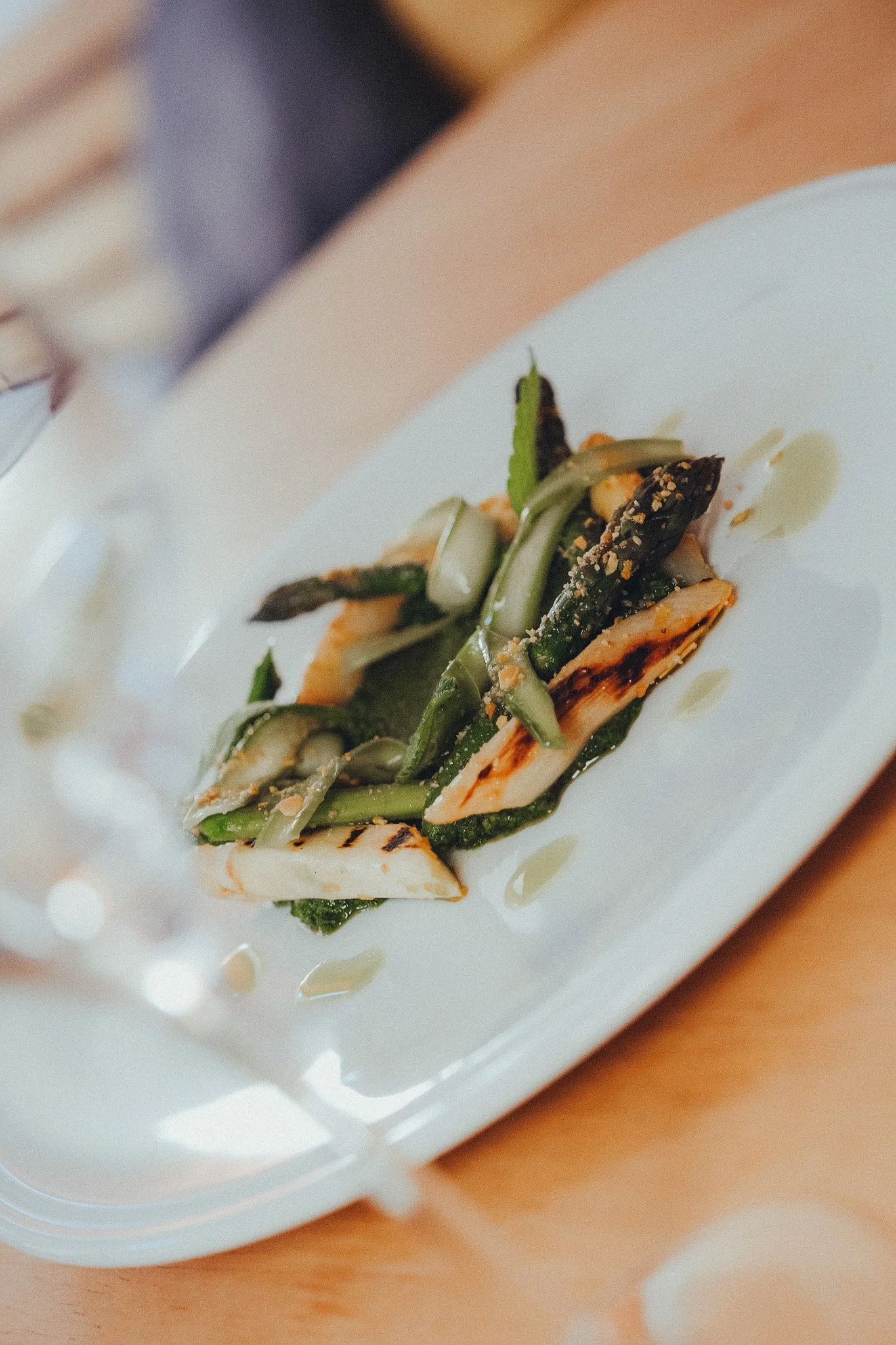 Close-up of a white rectangular plate with grilled chicken and sautéed green vegetables, topped with crushed nuts and drizzled with sauce, on a wooden table.