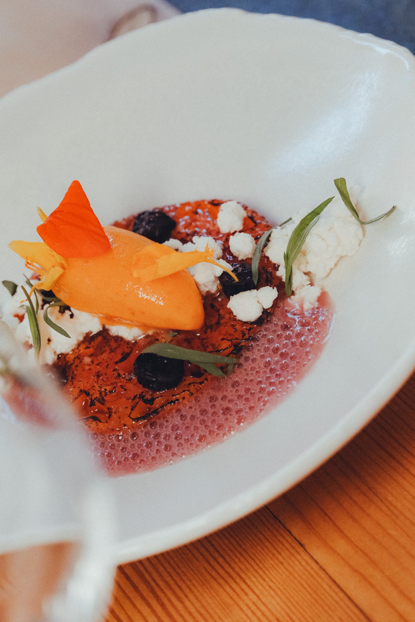 A close-up view of a dessert with whipped cream, berries, orange and green garnishes, served in a white bowl with a pink foam or sauce at the bottom, placed on a wooden table.