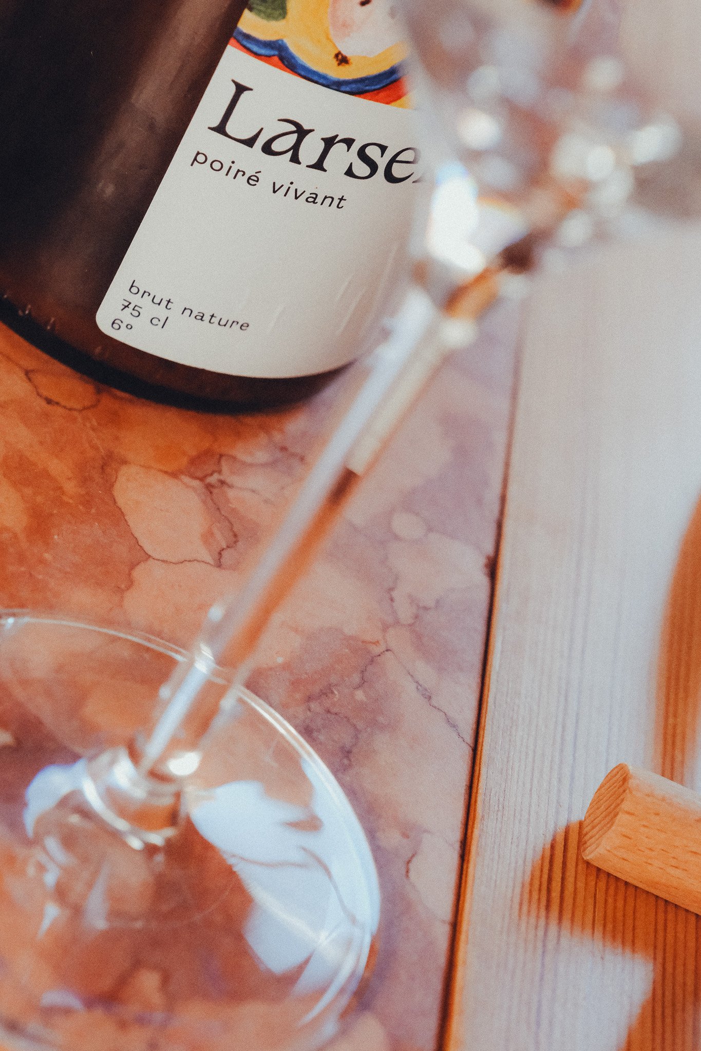 Close-up of a wine bottle label that reads 'Larse, Poiré Vivant, brut nature, 75 cl, 6°'. The background includes a glass and some wooden surface, with part of a white and orange wall.