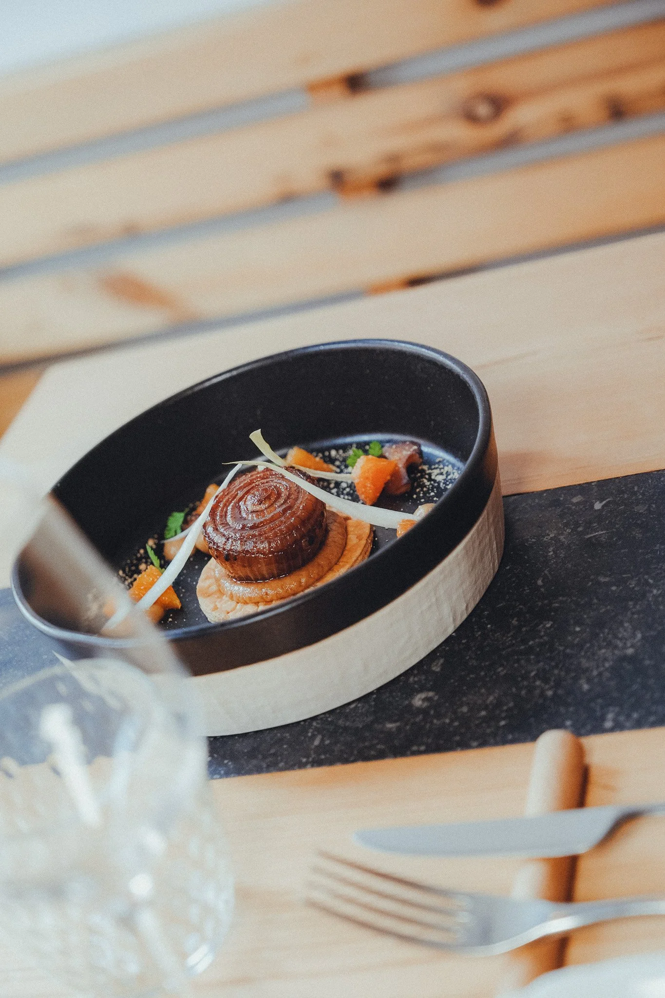 A black bowl with a wood base containing a lobster tail, garnished with vegetables and herbs, on a wooden tray on a dark countertop.