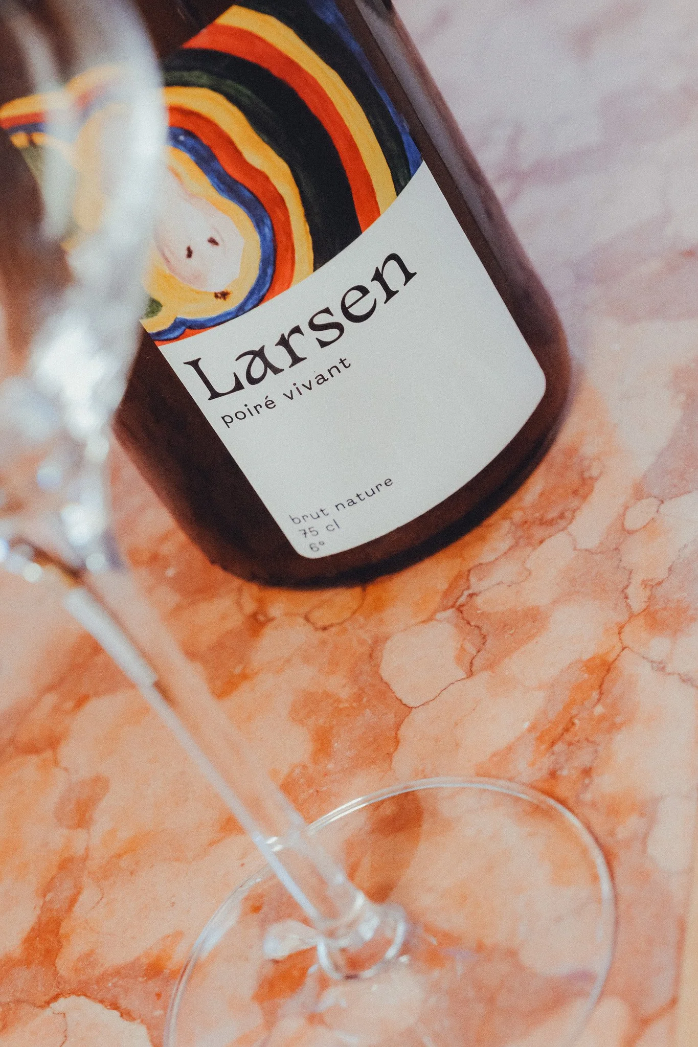 A bottle of Lamser brut nature sparkling wine with a colorful abstract label, placed on a pinkish-orange marble surface.