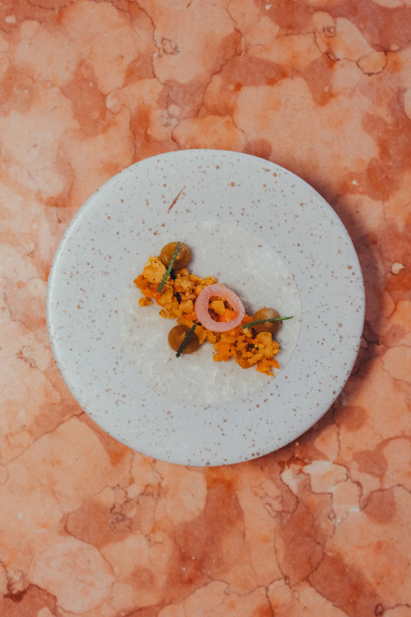 Close-up of a white plate with orange-colored food garnished with green herbs, red onion slice, and peas, placed on a pink marble surface.