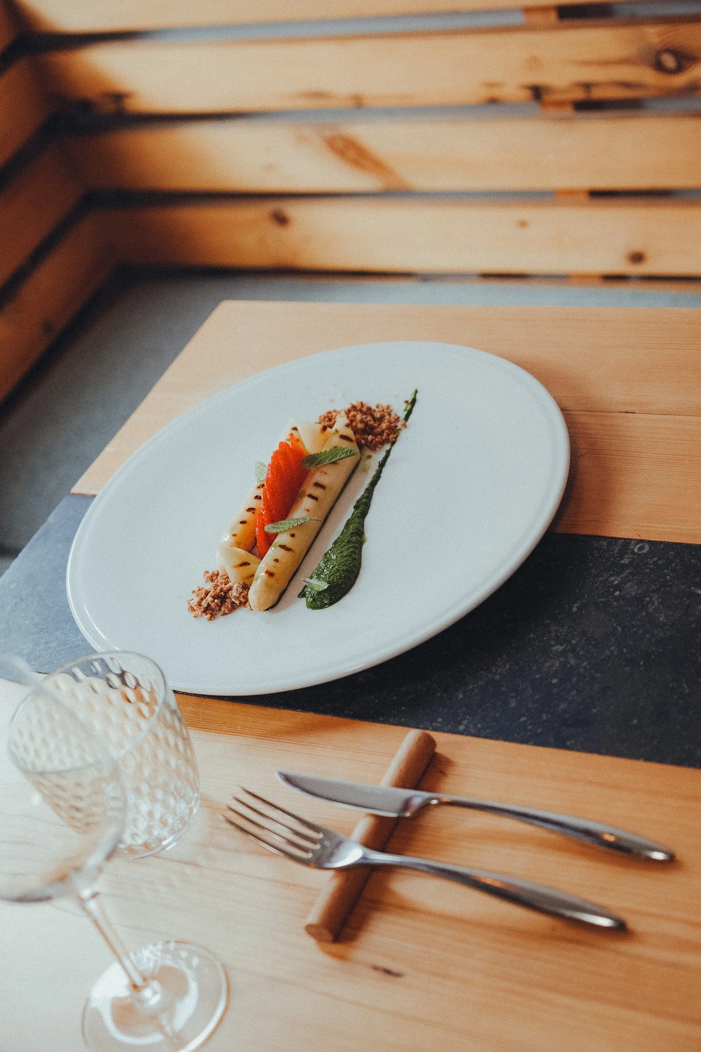 A white plate with grilled vegetables and crumbs on a wooden table, set with a fork, knife, and a textured glass, in a cozy dining setting.