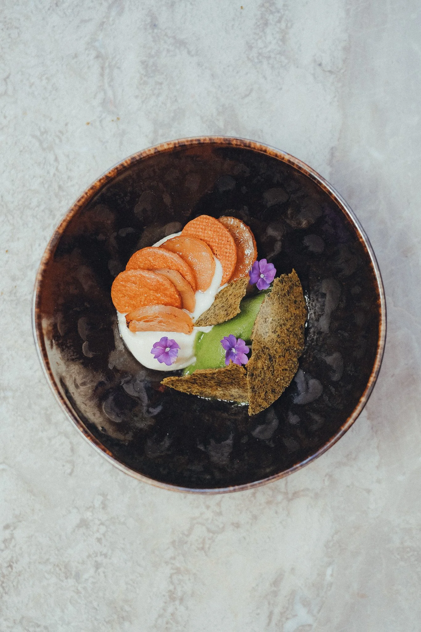 Dessert with whipped cream topped with orange and brown wafer cookies, green fruit slice, purple edible flowers, and crispy green garnish in a dark bowl, placed on a light textured surface.