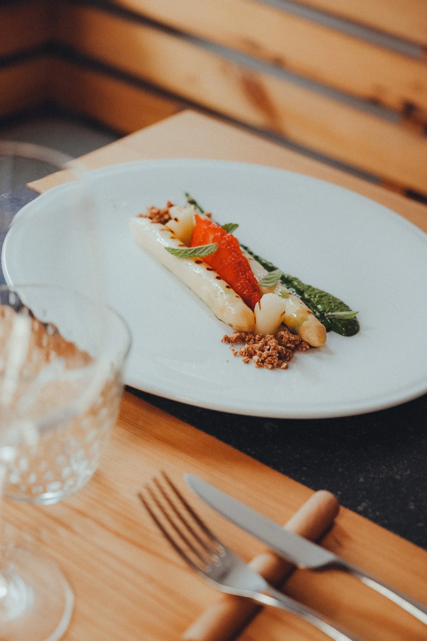 A plated dish featuring a variety of artistic food arrangements, including sliced vegetables, herbs, and a sauce, on a white plate set on a wooden table with cutlery.