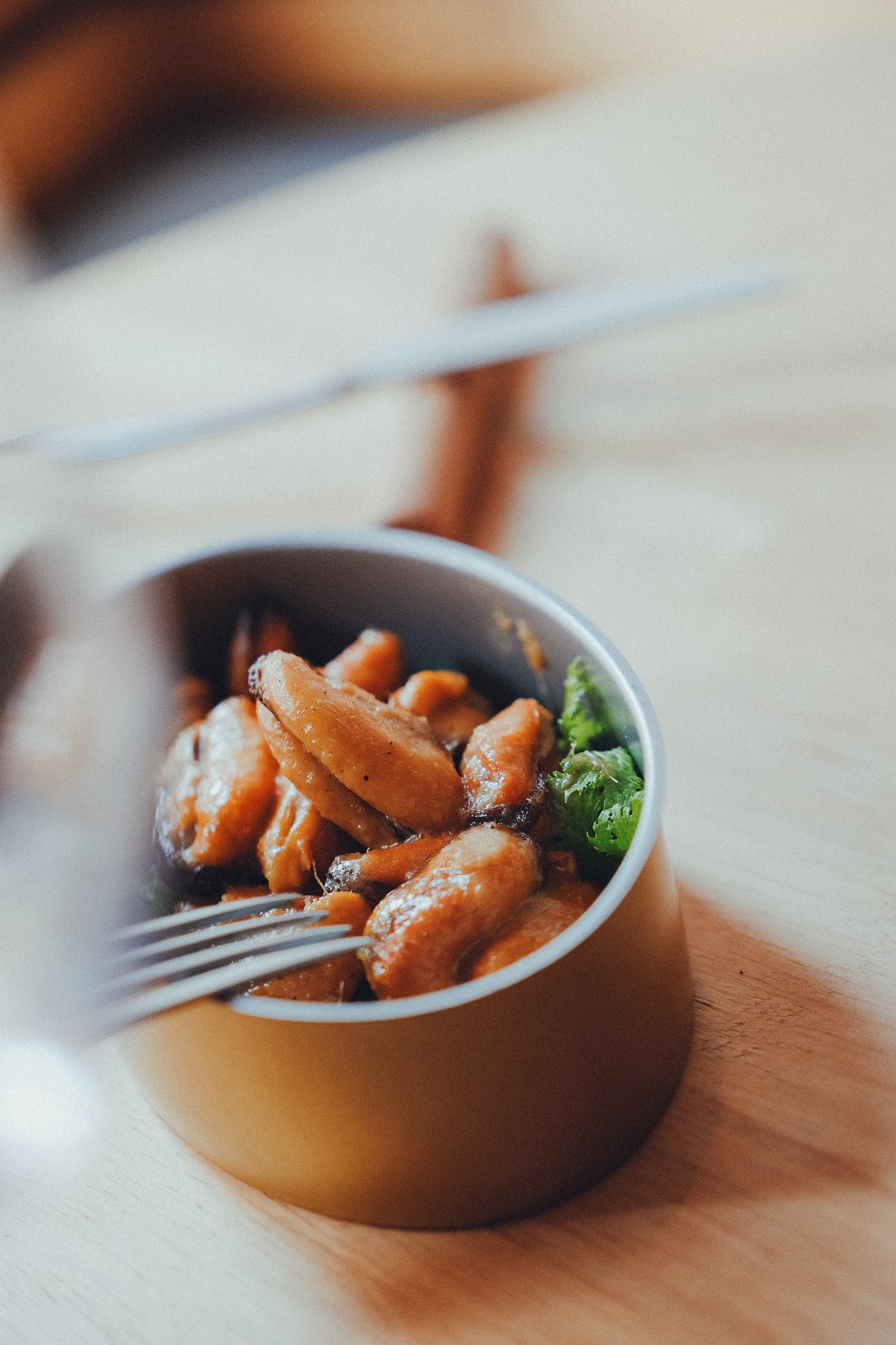 A bowl of cooked chicken pieces with broccoli on a wooden surface, with a fork and spoon nearby.