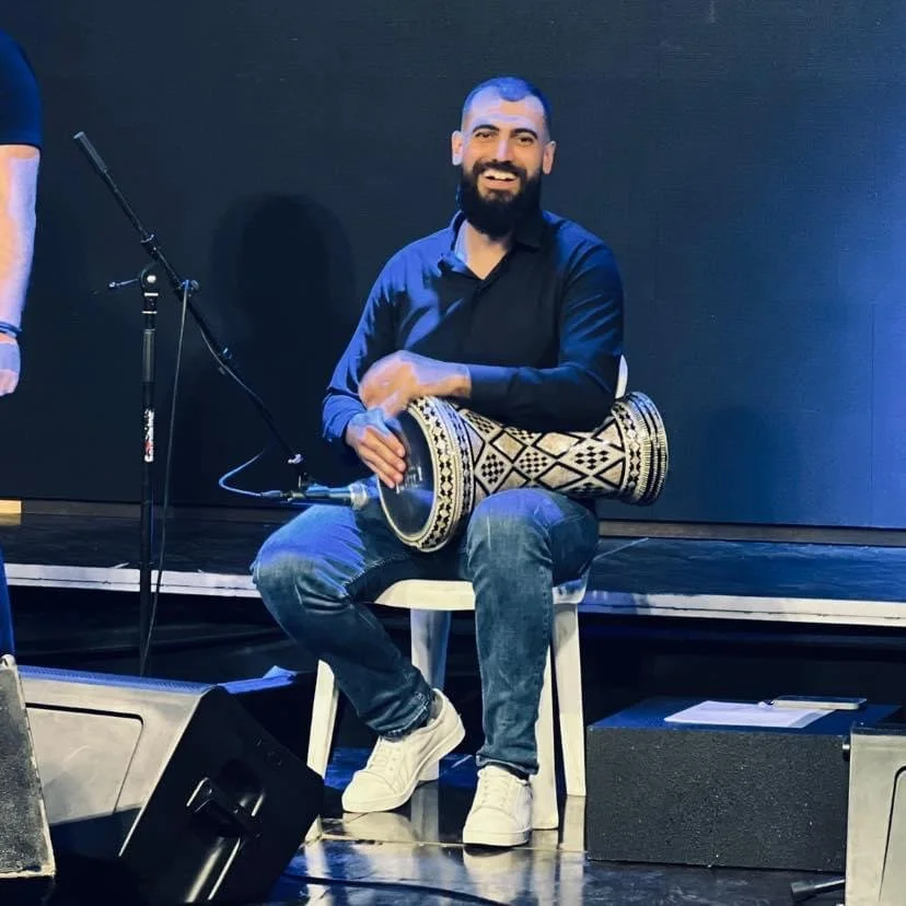 A man with a beard sitting on a chair, smiling, holding a traditional drum with a decorative pattern, on a stage with a black background.