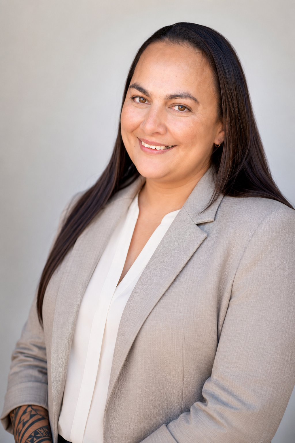 A professional woman with long dark hair, wearing a beige blazer over a white blouse, smiling at the camera against a plain light background.