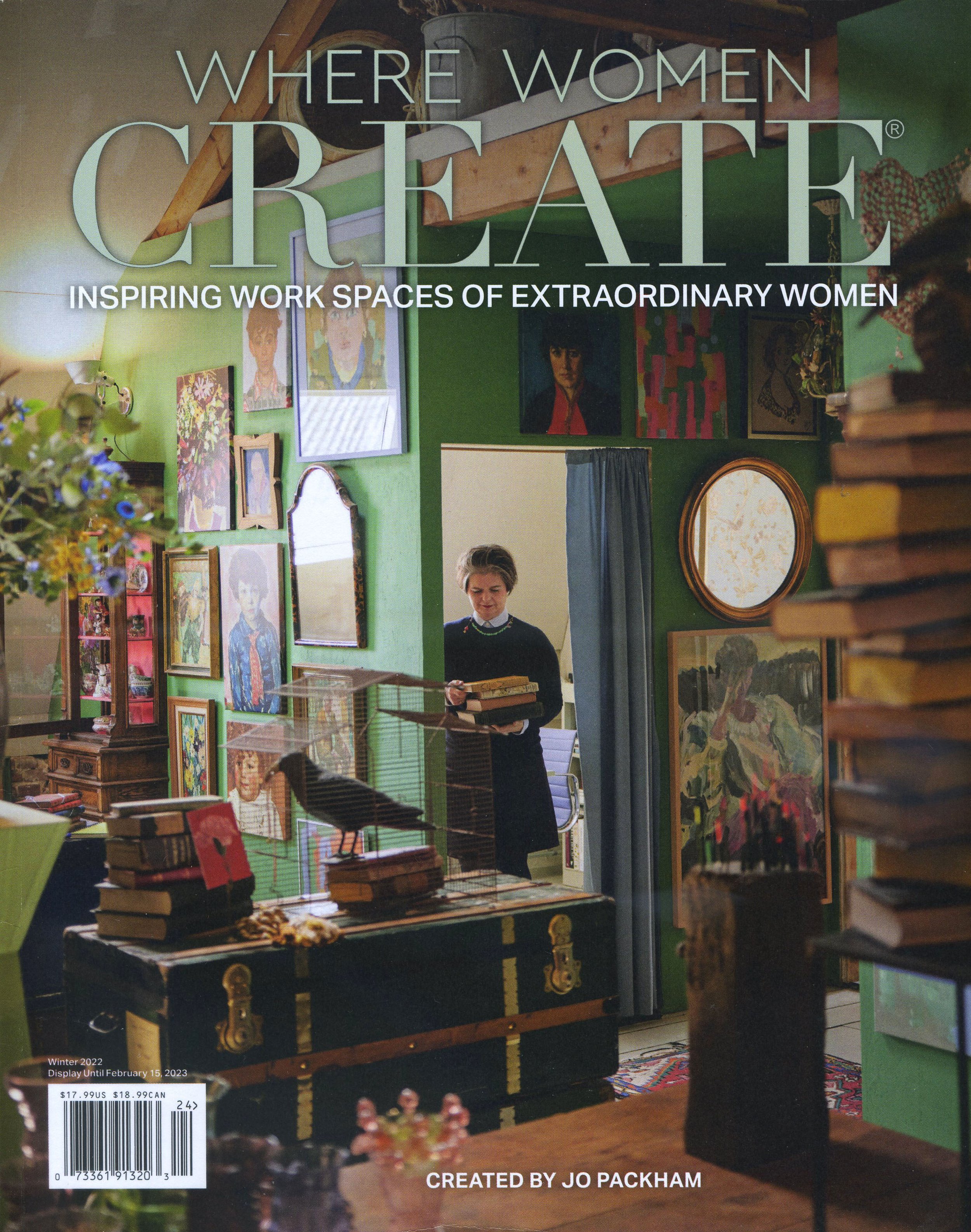 Inside view of an art studio or gallery with various paintings on the green walls, a woman holding a book in front of a mirror, and a black cabinet with gold accents in the foreground. There is a large mirror on the wall, and a colorful rug on the floor.