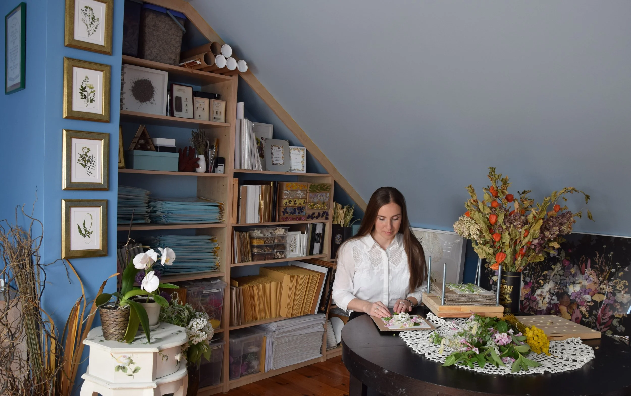 Anastasia Kovaleva working with dried flowers in her studio, surrounded by flower arrangements and plants, in a room with blue walls and wooden shelves