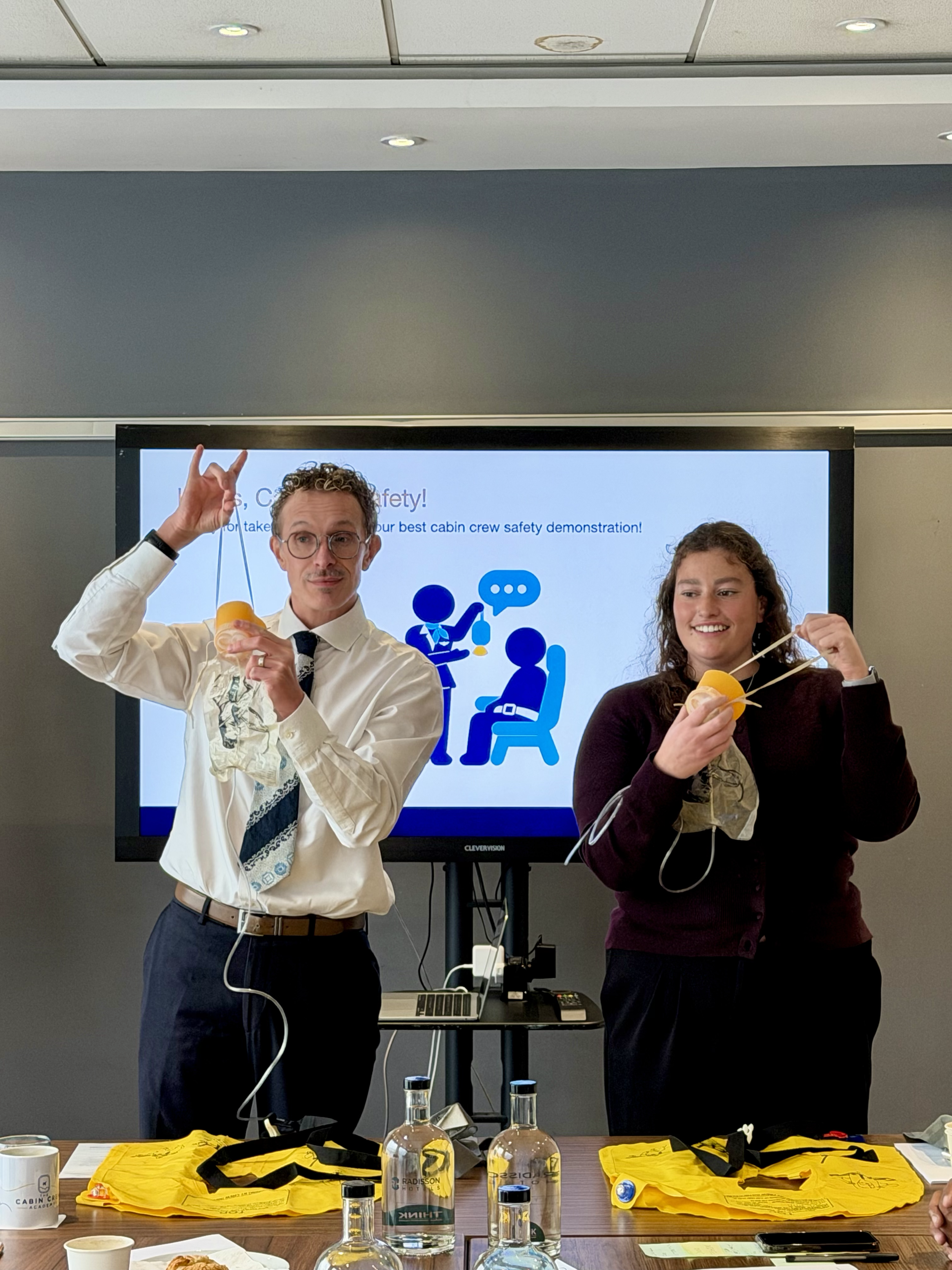 Two presenters demonstrate an airplane safety presentation using oxygen masks in front of a screen.