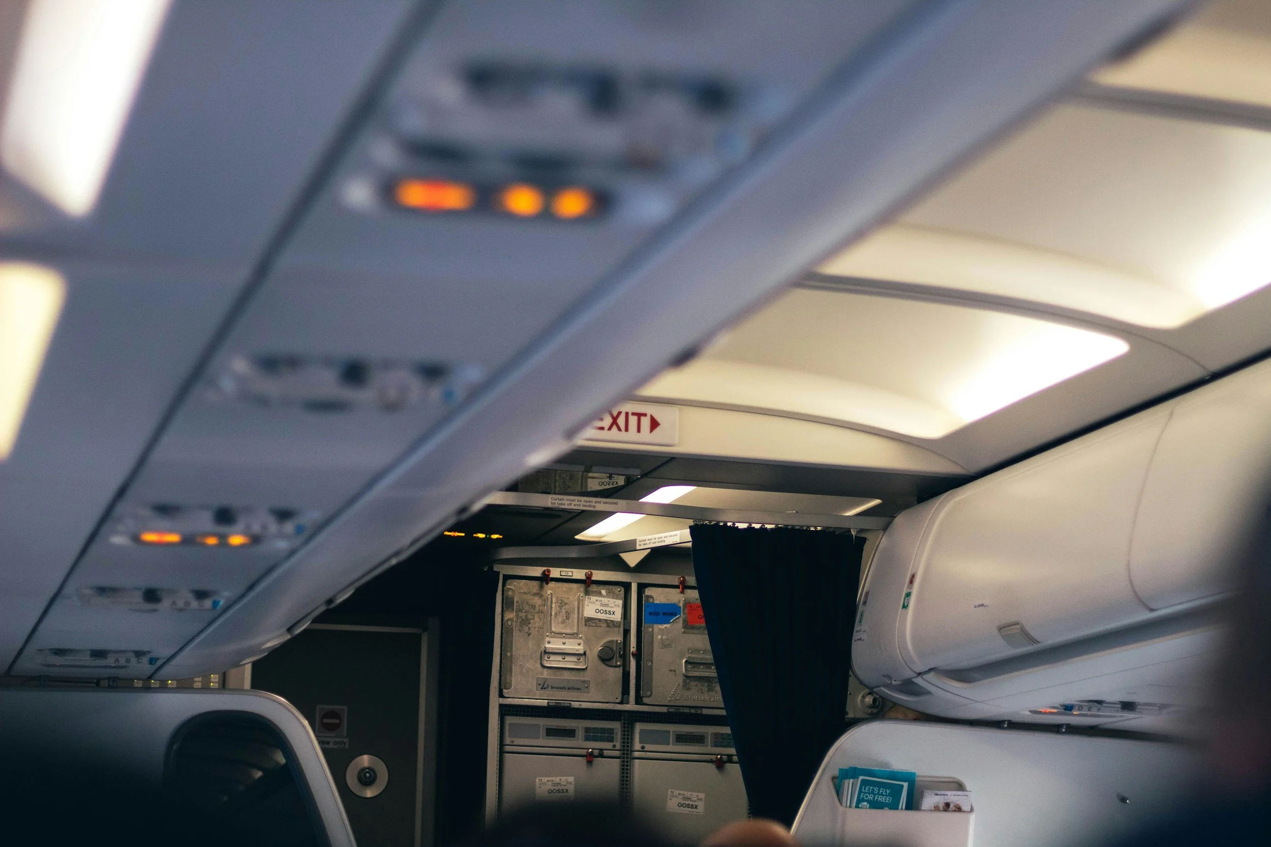 Inside the cabin of an airplane showing overhead controls, a safety sign, and part of a seat with a window.