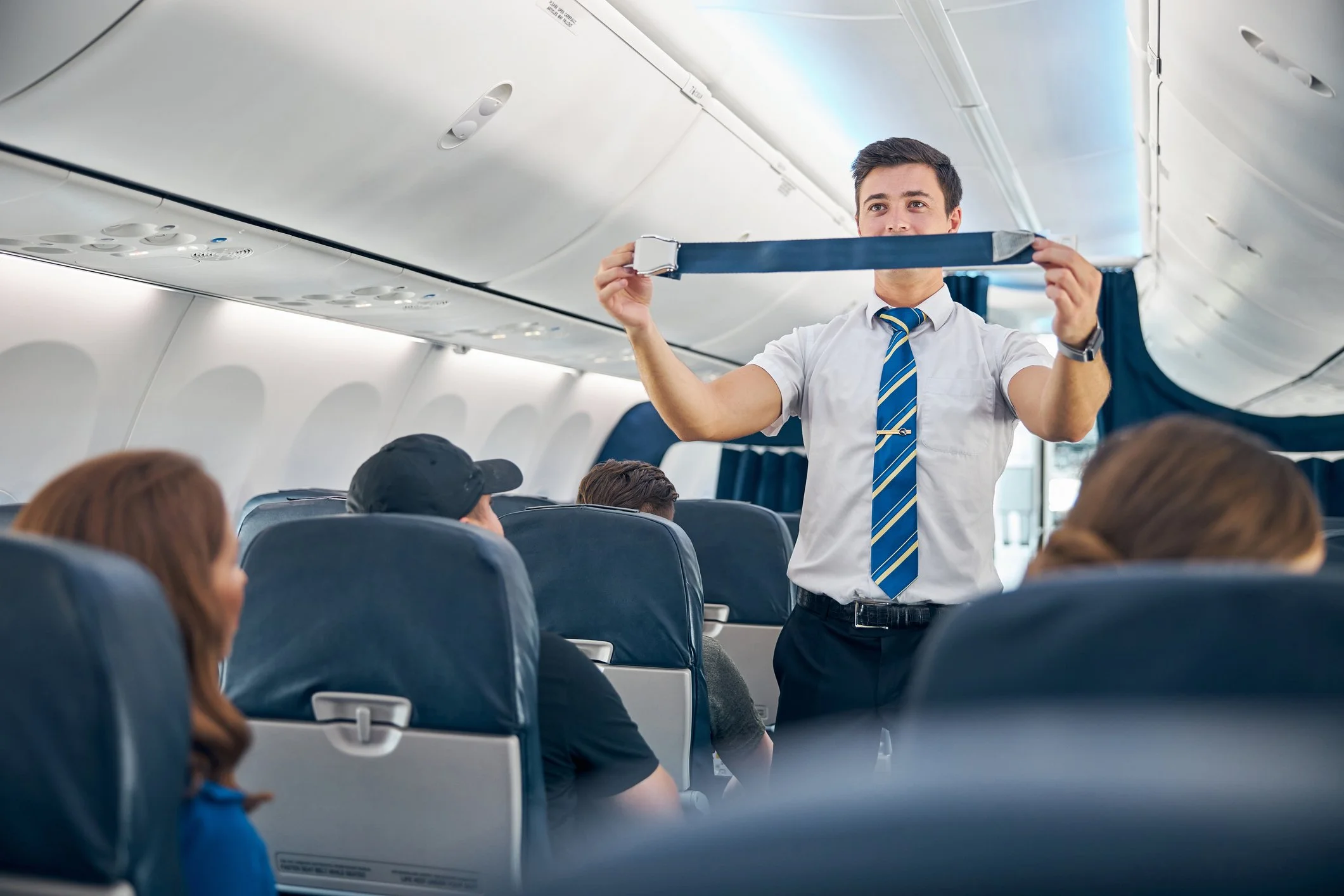A male flight attendant demonstrating safety procedures on an airplane, holding a seat belt in the cabin with passengers seated around him.