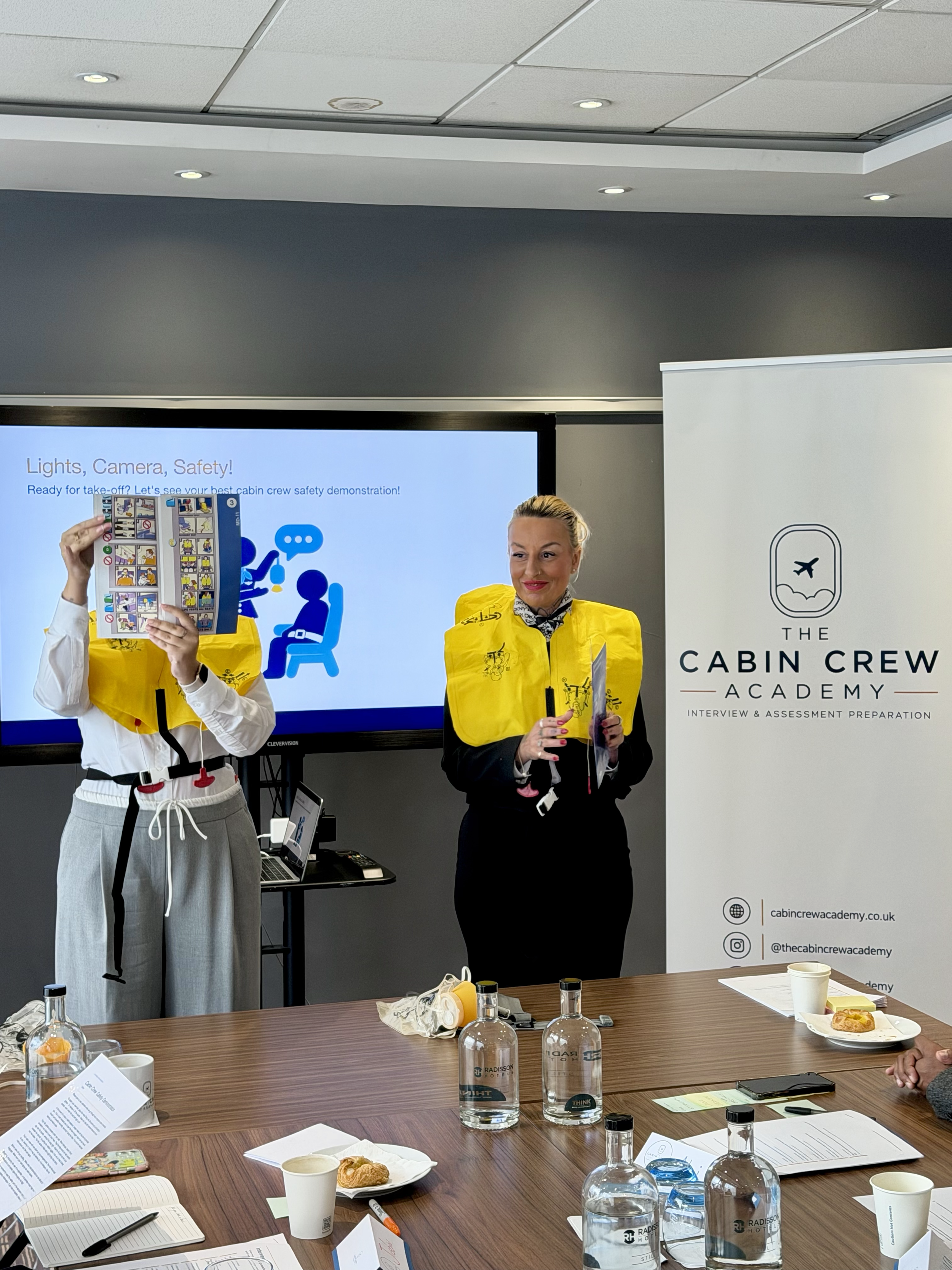 Cabin crew training session with two instructors demonstrating airline safety procedures in front of a presentation screen and Cabin Crew Academy banner.