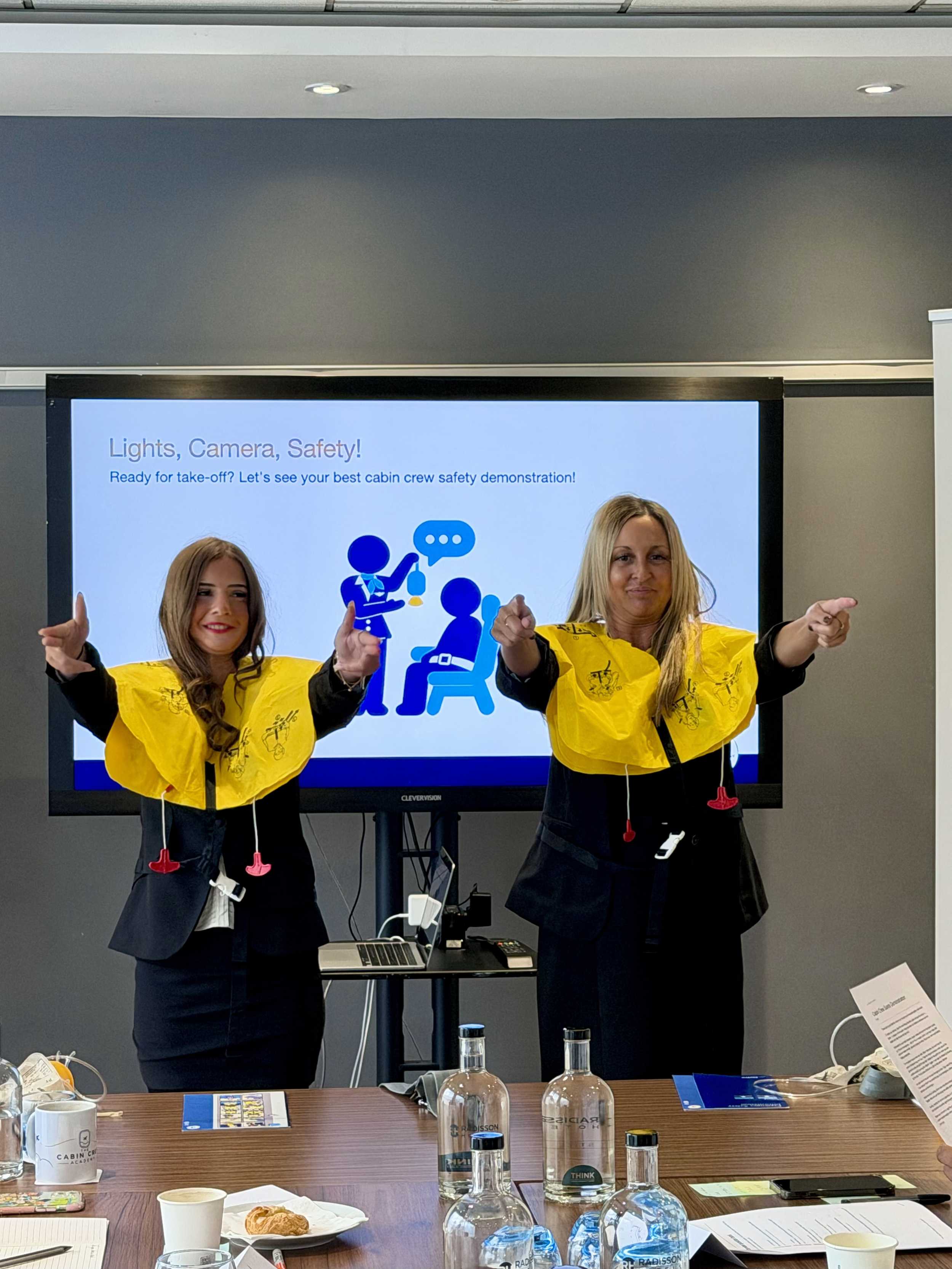 Two women in yellow airplane safety vests demonstrate cabin crew procedures in front of a presentation screen.