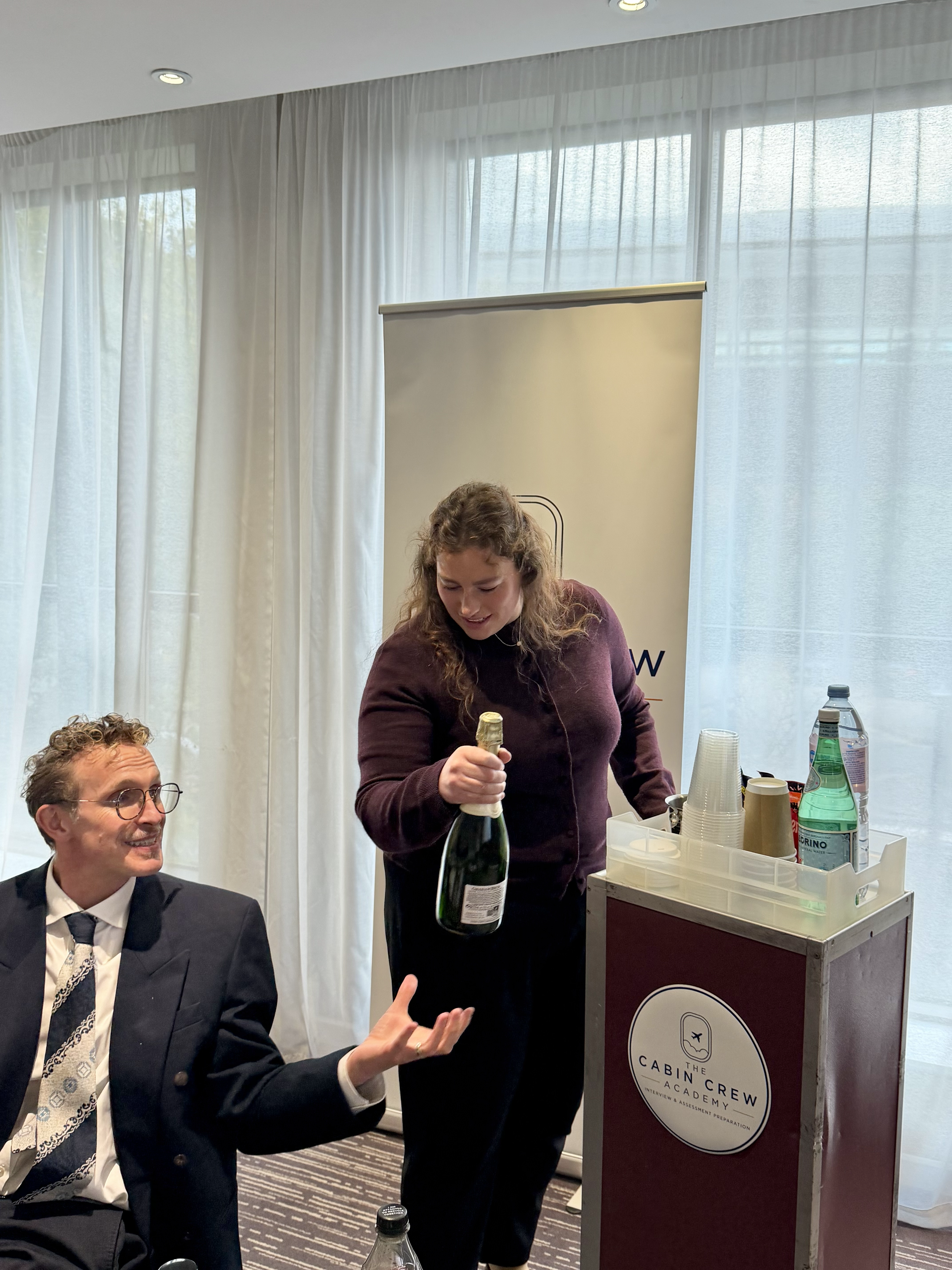 Woman opening a champagne bottle beside a seated man at a Cabin Crew Academy event